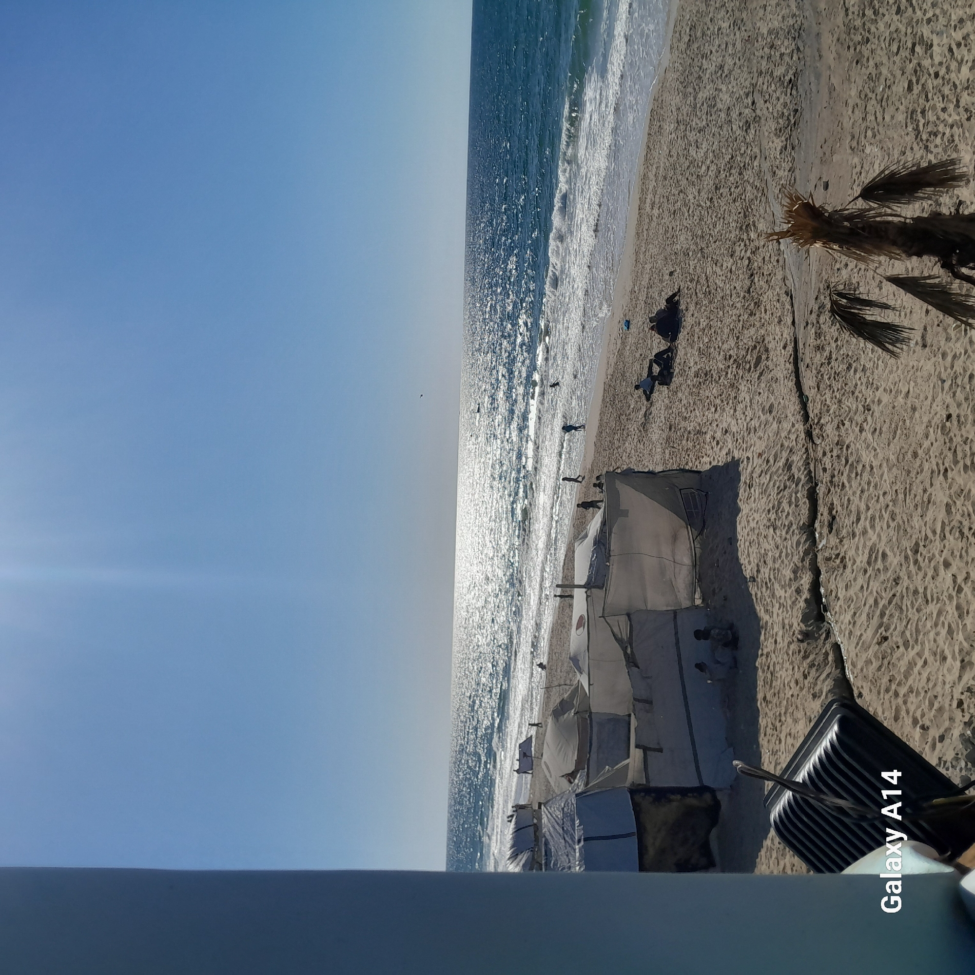 The image shows a beach in Unknown, Unknown. The scene includes the ocean, a sandy beach with tents, and several people. The sky is clear and blue. A suitcase is visible in the foreground.