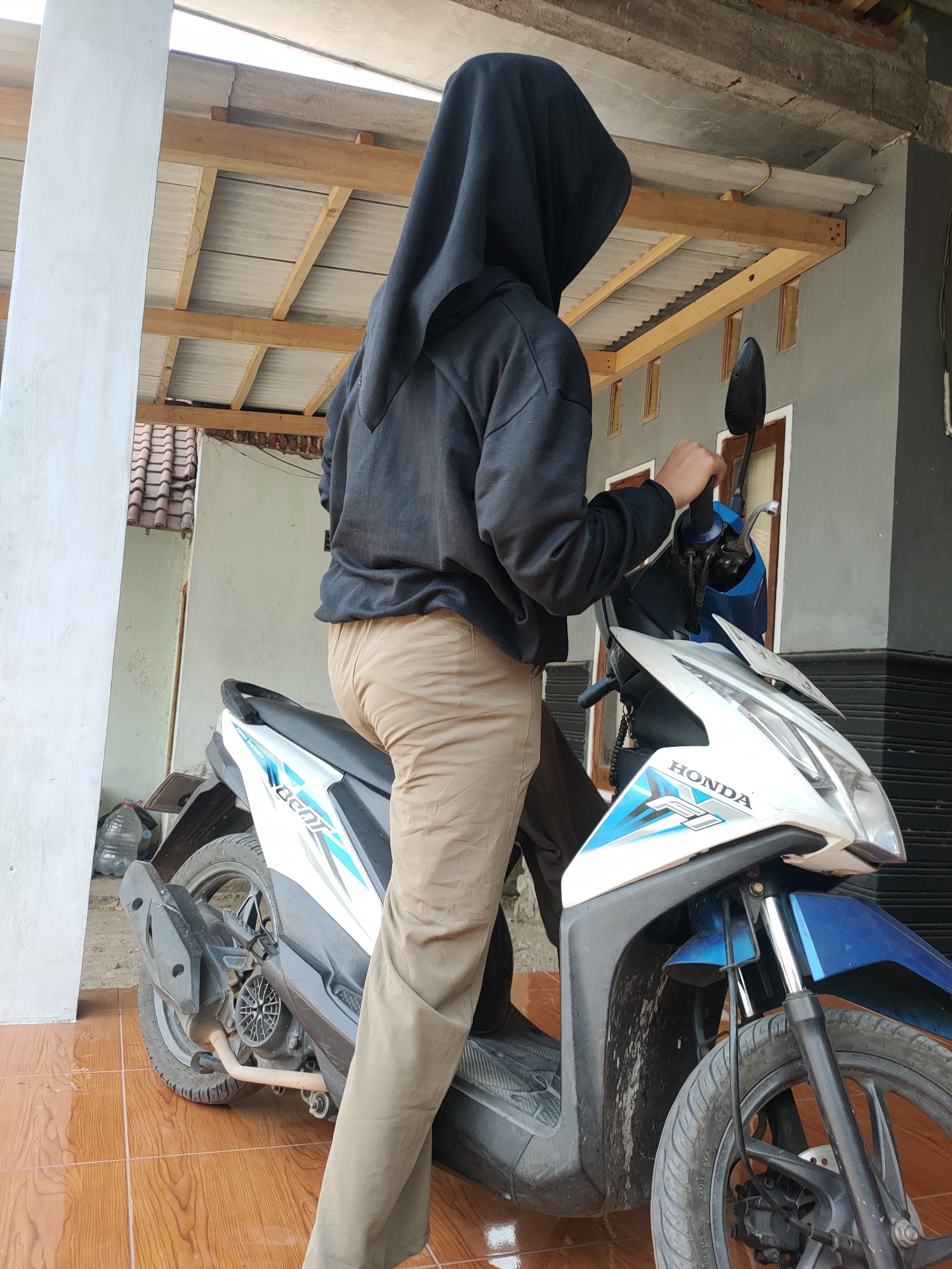 A person wearing a black hijab and a black hoodie is positioned on a white and blue Honda Beat scooter. They are wearing khaki pants and are holding onto the scooter's handlebars. The scooter is parked on a brown tiled floor, likely outdoors, with a structure overhead supported by wooden beams and corrugated roofing. In the background, a gray wall with a window and a section of a tiled roof are visible. A plastic water bottle is on the ground to the left of the scooter. The scene appears to be in a residential area in Kanigoro, Indonesia, possibly a yard or a covered parking space.