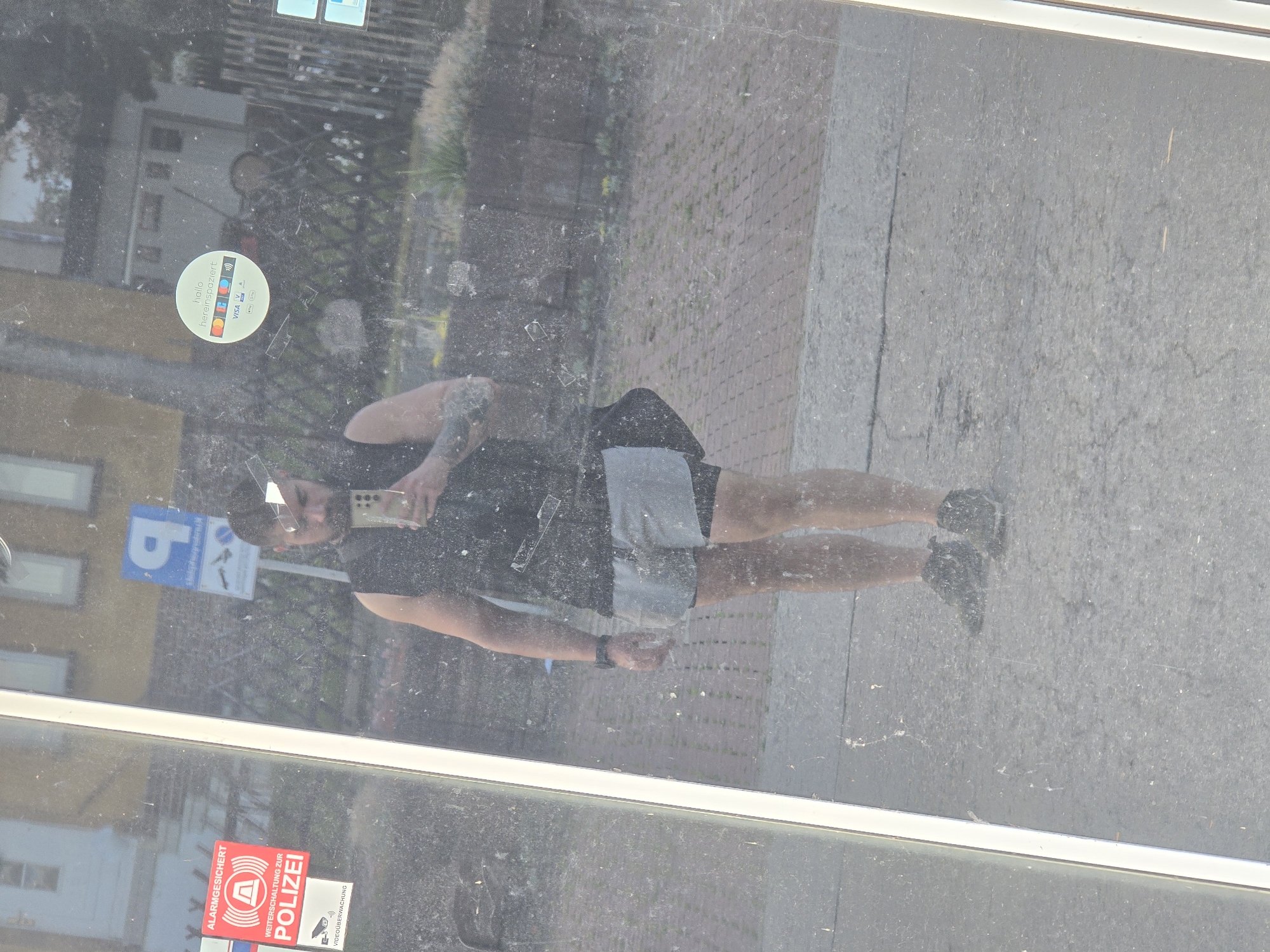 A man is seen reflected in a dirty glass surface, taking a mirror selfie. He is wearing a black sleeveless top, grey athletic shorts over black compression shorts, and dark sneakers. He is also wearing a dark watch on his left wrist and holding a smartphone in his right hand. Behind him, the reflection shows a paved ground, brickwork, a sidewalk with some plants, and a fence. There are several stickers visible on the glass surface in front of the reflection. One is a red and white sticker with German text that appears to be an alarm system warning. Another is a circular sticker with German text and various payment card logos. A blue parking sign is also reflected. The overall environment suggests an urban setting.