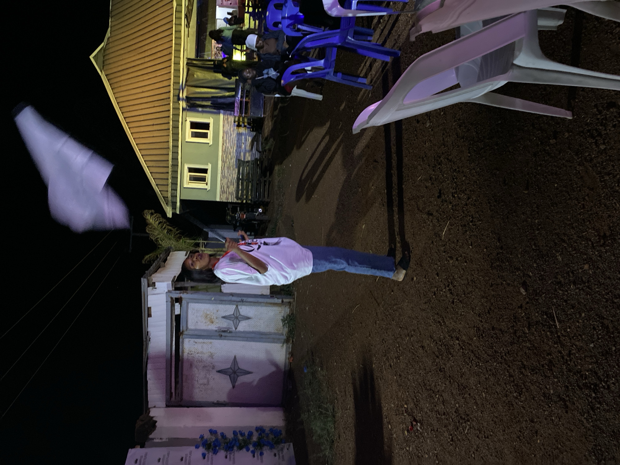 At night in Durimi, Nigeria, a person in a white t-shirt and jeans stands next to a gray building with a metal gate adorned with star-shaped decorations. They hold green foliage. A tombstone with blue flowers sits at the building's base. In the background, a gathering of people sit on blue plastic chairs. A building with a corrugated metal roof is visible, with light emanating from its windows. White plastic chairs are stacked nearby. The ground is dark and appears to be dirt or gravel.