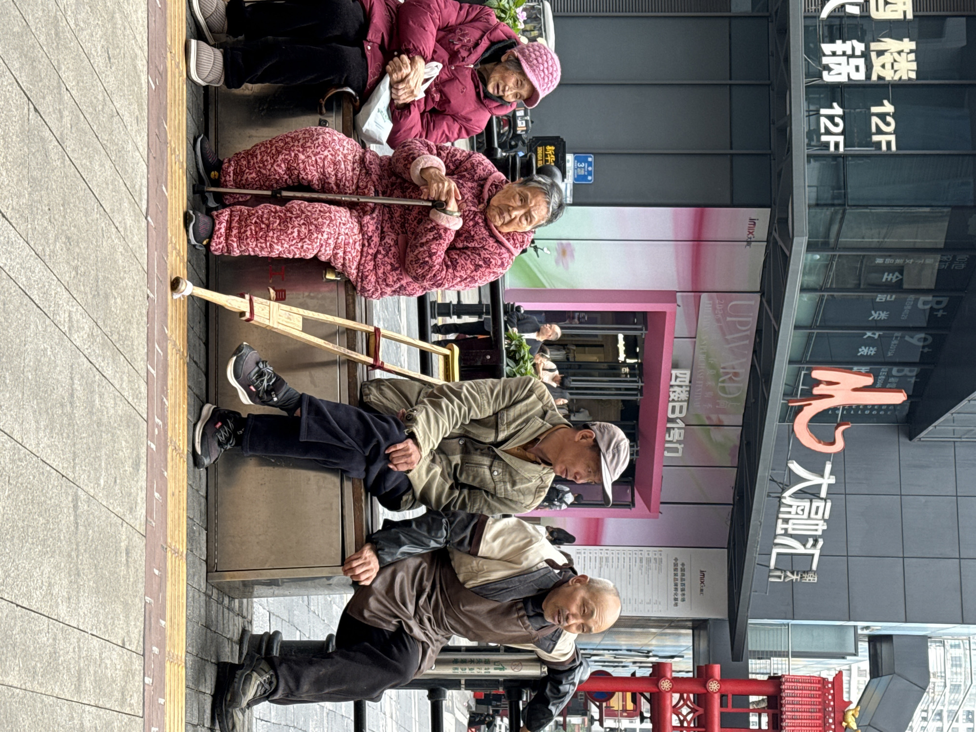 Four elderly people are sitting outside a building on a sidewalk in the Yuzhong District of China. Two women are sitting on a bench, one wearing a red quilted jacket and a pink knit hat, and the other in a pink patterned quilted jacket and using a walking stick. Two men are sitting on a lower level bench in front of them. The first man is wearing a brown jacket and the second is wearing a green jacket and a cap. Behind the people is a building with signs in Chinese characters. A red and gold decorative structure is in front of the building. A tactile paving strip lines the edge of the sidewalk.