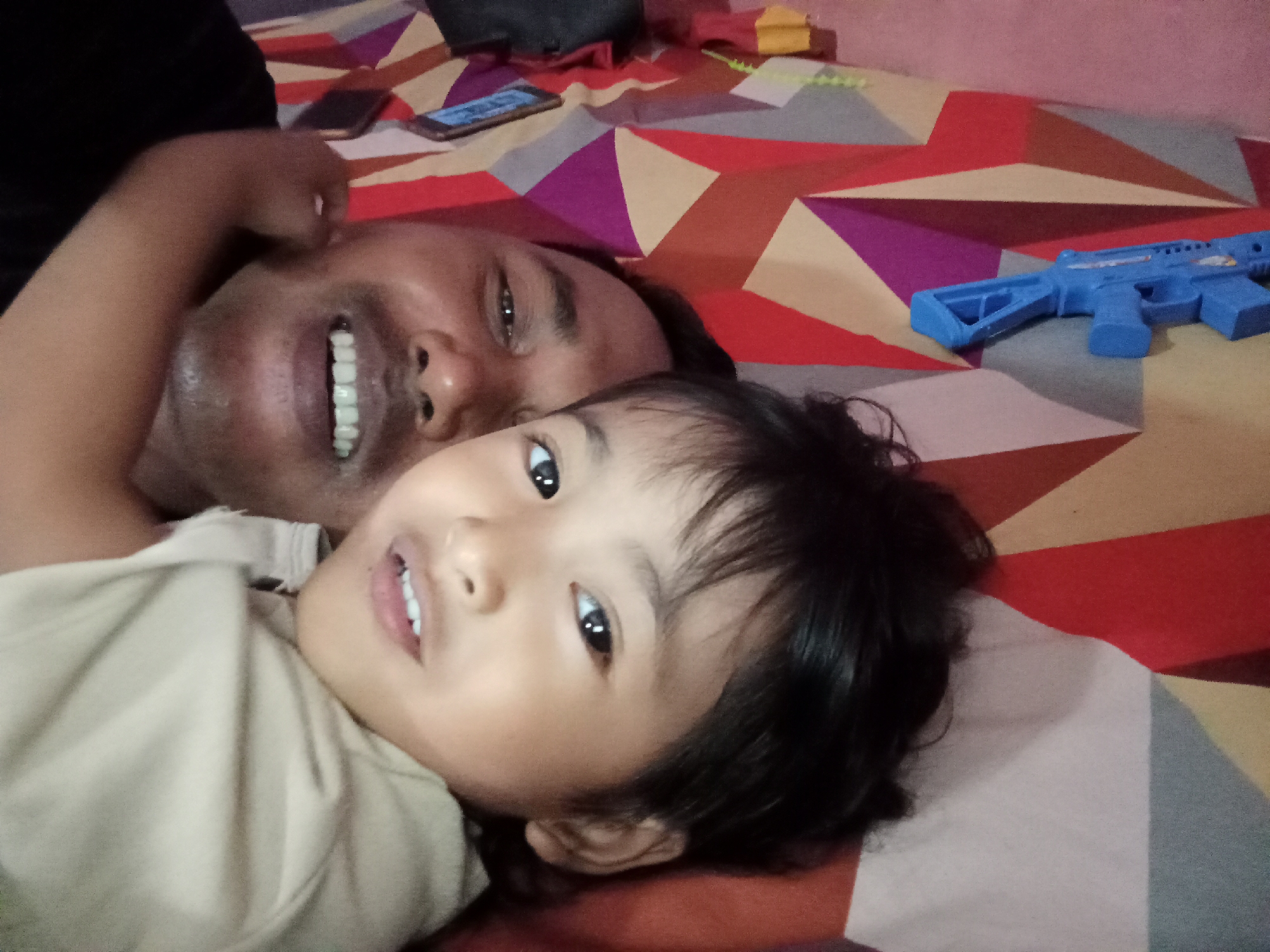 The image shows a close-up, high-angle shot of a man and a young child lying on a bed with a vibrantly patterned bedsheet. The man is on the left, with his arm around the child and his face turned towards the camera, smiling broadly, showing his teeth. The child is positioned in front of the man, also facing the camera with a gentle smile, and their eyes are wide open.
The bedsheet has a geometric pattern of red, purple, yellow, and grey triangles and quadrilaterals. On the bed, to the right of the people, a blue toy gun is visible, along with what appear to be cell phones. The background is blurred, suggesting a domestic setting, possibly a bedroom. The lighting is soft, indicating an indoor environment.
The image quality is somewhat low, with a slight blurriness and graininess throughout. The perspective suggests the photo was taken by someone lying down, likely the man or child. The presence of the toy gun and cell phones indicates a contemporary setting. While the image itself does not contain explicit clues to its specific location within Bloro, Indonesia, such domestic scenes are common in households there. The overall impression is one of intimacy and shared leisure time between the man and child.