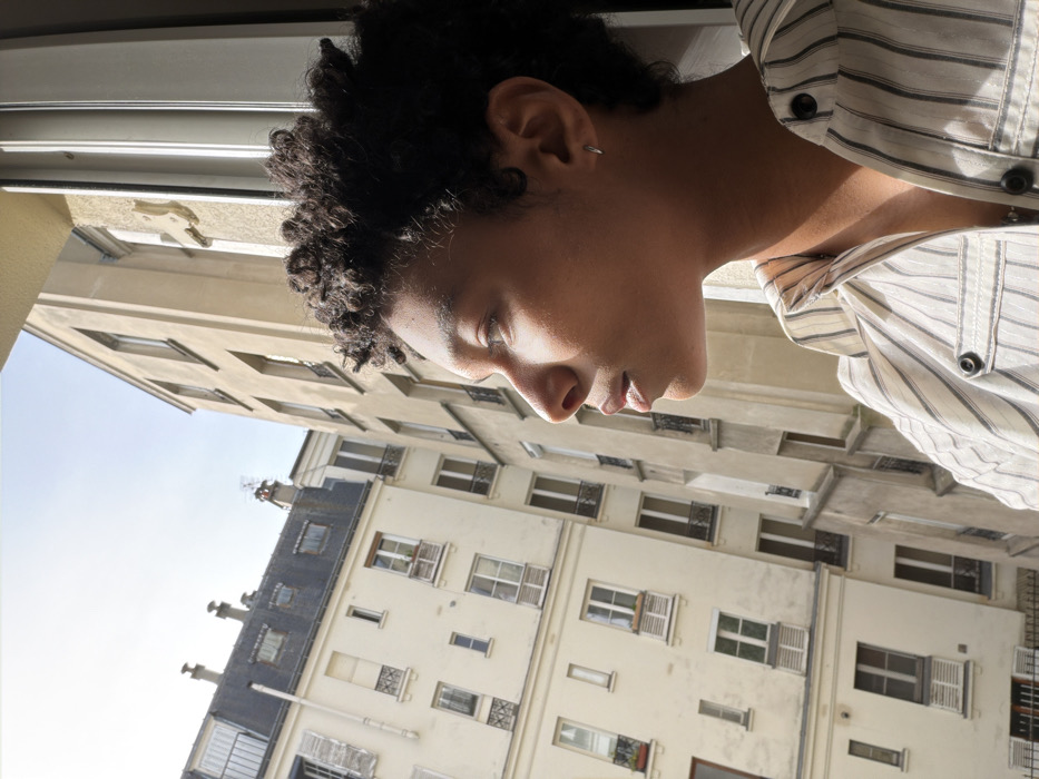 The image depicts a close-up, profile view of a person looking out from an open window in the city of Paris, France. The individual, positioned on the right side of the frame, has dark, tightly curled hair and is looking towards the left. Sunlight illuminates their right cheek and jawline. They wear a light-colored, vertically striped button-up shirt with dark buttons, and a small silver hoop earring is visible in their right ear.
Beyond the window, a typical Parisian urban landscape is visible under a clear, bright sky. Two multi-story buildings with light-colored facades dominate the background. The building on the left features numerous rectangular windows, some with closed shutters, and several small balconies with wrought-iron railings. Its upper section includes a dark grey mansard roof with dormer windows and chimneys, along with a visible antenna. The building directly opposite the window, on the right, appears to be a similar Haussmannian-style structure, partially visible, also with multiple windows. The scene is brightly lit, suggesting daytime.