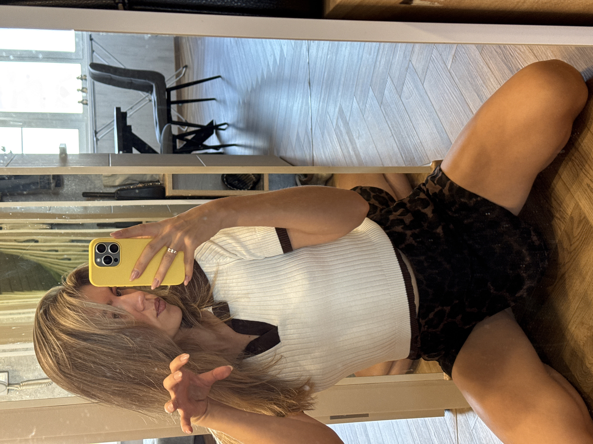 A woman is seen in a mirror, taking a selfie. She is wearing a white, short-sleeved top with brown trim and brown shorts. The background appears to be an interior space with a wooden floor, a chair, and a window. The location and city are unknown.