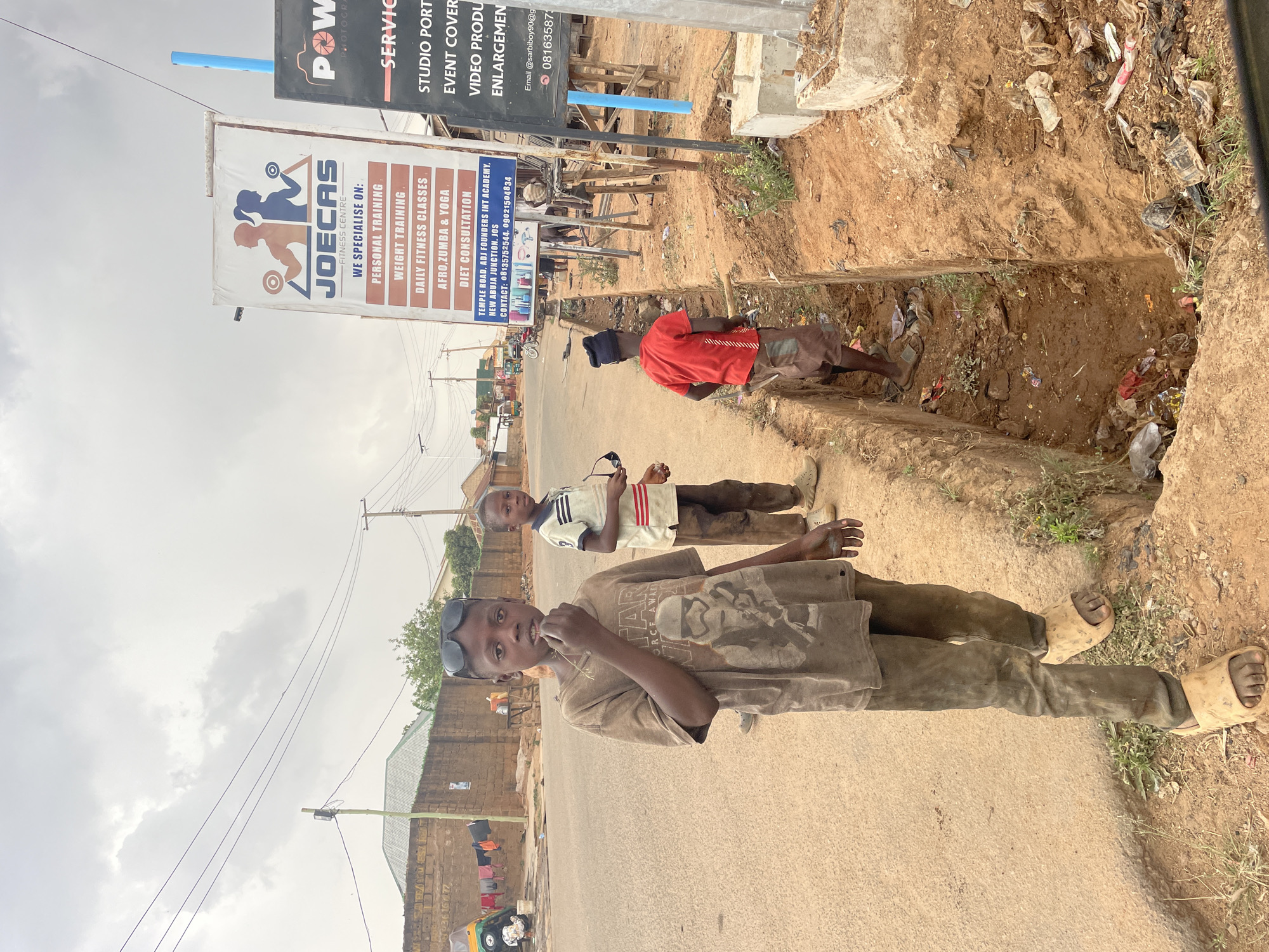 Three boys stand on a street in Jos, Nigeria. The foreground boy wears a brown shirt with a graphic, brown pants, and beige sandals. He also has sunglasses perched on his head and is holding something in his mouth. The middle boy wears a striped shirt with red accents and brown pants. He is holding sunglasses. The background boy is wearing a red shirt and dark shorts. They stand near an open, trash-filled drainage ditch. In the background, a sign reads "JOECAS FITNESS CENTRE." Next to it is a sign that advertises photography services. The street is asphalt and there are buildings, power lines, and parked vehicles further down the street.