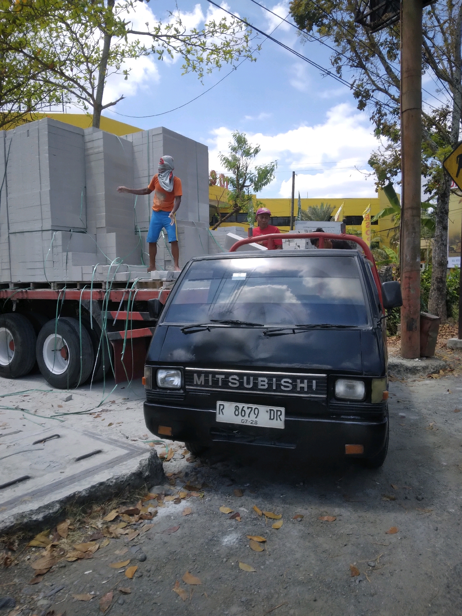 The image depicts a street scene in Sokaraja, Indonesia, with a parked black Mitsubishi pickup truck in the foreground. The truck has a license plate reading "R 8679 DR" and "07-28". Behind the truck is a flatbed truck laden with stacks of light grey building blocks, secured with green straps. A man in an orange t-shirt and blue shorts, with his face covered by a grey cloth, stands on the building blocks and gestures with his right hand. Another man wearing a red shirt and a red cap is partially visible near the cab of the flatbed truck. The background features a yellow building, trees, and a utility pole with a traffic sign. The ground is a mix of dirt, gravel, and fallen leaves.