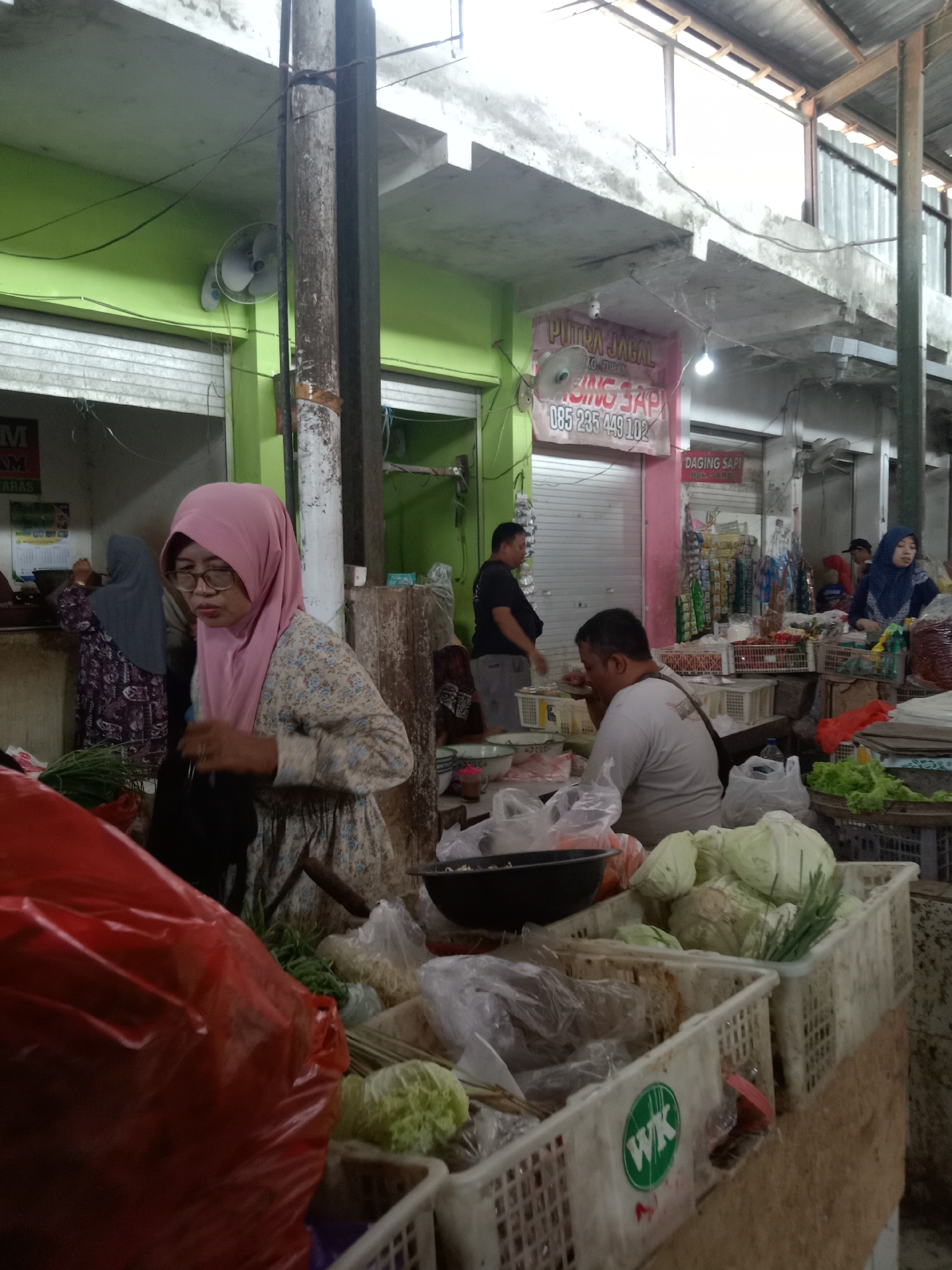 This is a mid-angle, indoor shot of a bustling market scene in Sugihwaras, Indonesia. The market appears to be a wet market, with fresh produce prominently displayed. In the foreground, a woman wearing a pink headscarf and glasses is focused on a transaction, holding a dark bag. She is standing behind a display of red plastic bags filled with what appears to be produce, and other vegetables like scallions and cabbage are visible in white plastic crates.
The environment is characterized by an open-air market structure with a corrugated metal roof supported by wooden beams and concrete pillars. The walls of the market stalls are painted in vibrant colors, with lime green visible on the left and pink on the right. Signage is present, including a pink banner with Indonesian text that reads "PUTRA JAGAL" and a phone number "085 235 449 102". Another sign in red text reads "DAGING SAPI" (beef).
Several other individuals are present in the scene, including a man in a black t-shirt and grey pants standing near a stall, and another man with a light-colored shirt sitting at a table laden with produce. A woman in a blue headscarf is seen serving customers at a stall further back. The lighting is somewhat dim, suggesting it may be early morning or late afternoon, common times for such markets. The overall atmosphere is one of commercial activity and daily life in a local Indonesian market.