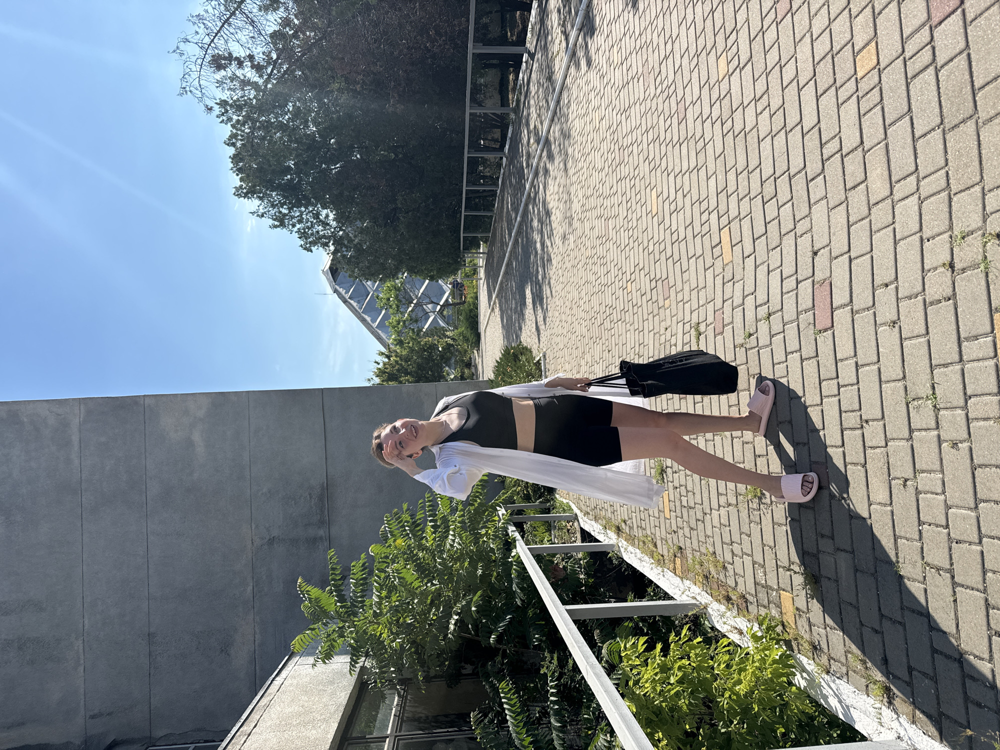 A young woman stands on a paved walkway outside on a sunny day, wearing a white open button-up shirt over a black sports bra and black shorts. She is holding a black tote bag in her right hand and has her left hand raised to her forehead, shielding her eyes from the sun. She is wearing pink slide sandals. To her left, a gray building with horizontal lines is visible, with a railing and lush green foliage alongside the walkway. In the background, trees and a modern glass-roofed structure with a geometric design are visible under a clear blue sky with sun glare. The walkway is made of interlocking brick pavers. The scene appears to be in a park or public outdoor space.