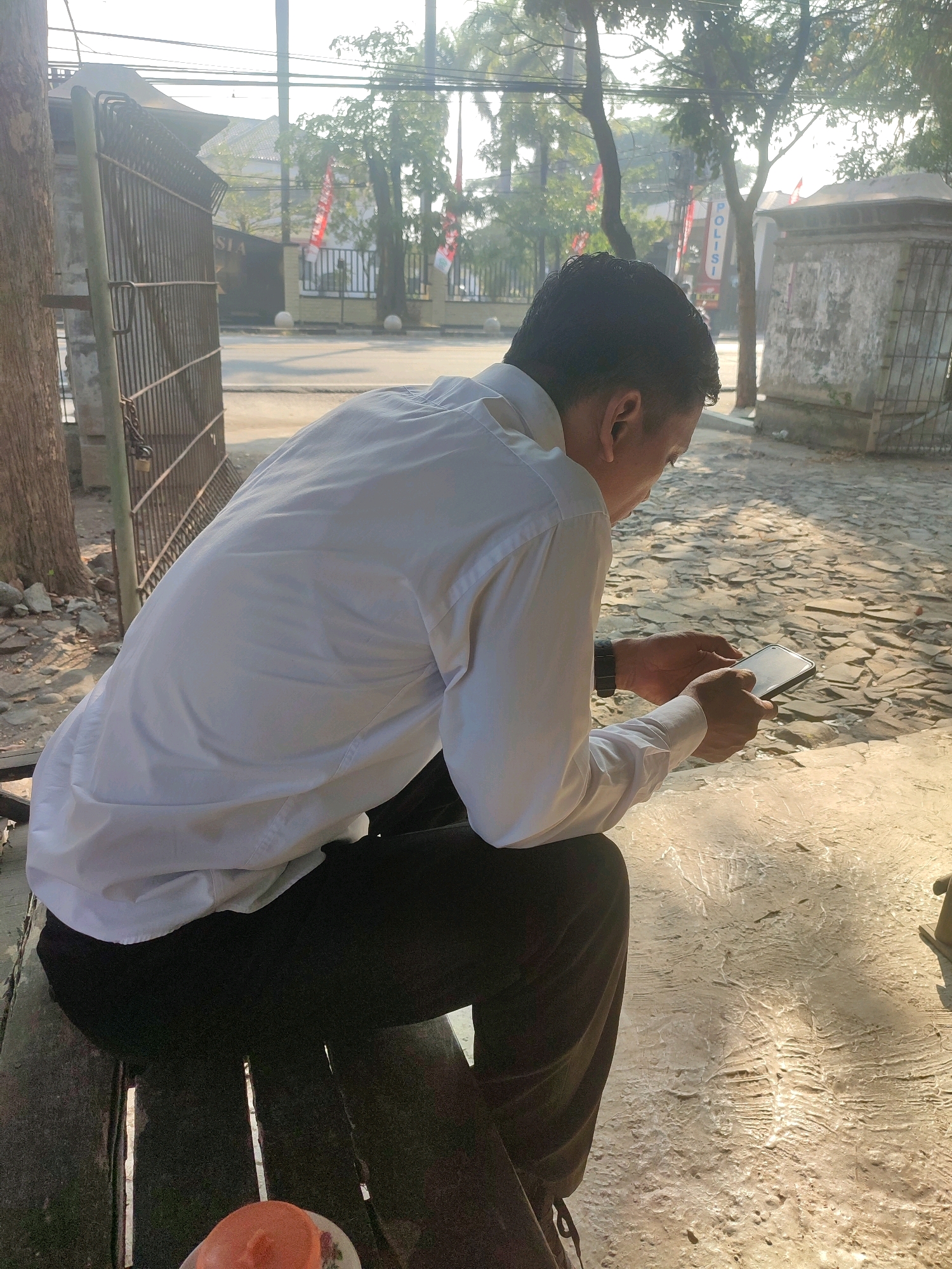 A person is sitting on a bench outdoors, wearing a white long-sleeved shirt and black pants, and looking at a smartphone. The person's back is to the camera. The environment appears to be a paved area with some stone-like textures on the ground. Behind the person, there is a metal gate, trees, and some buildings with flags. The overall lighting suggests it is daytime. Based on visual cues such as the architectural style and the presence of what appears to be the Indonesian flag and the word "POLISI" (police in Indonesian), this scene is likely located in Ringinanom, Indonesia.