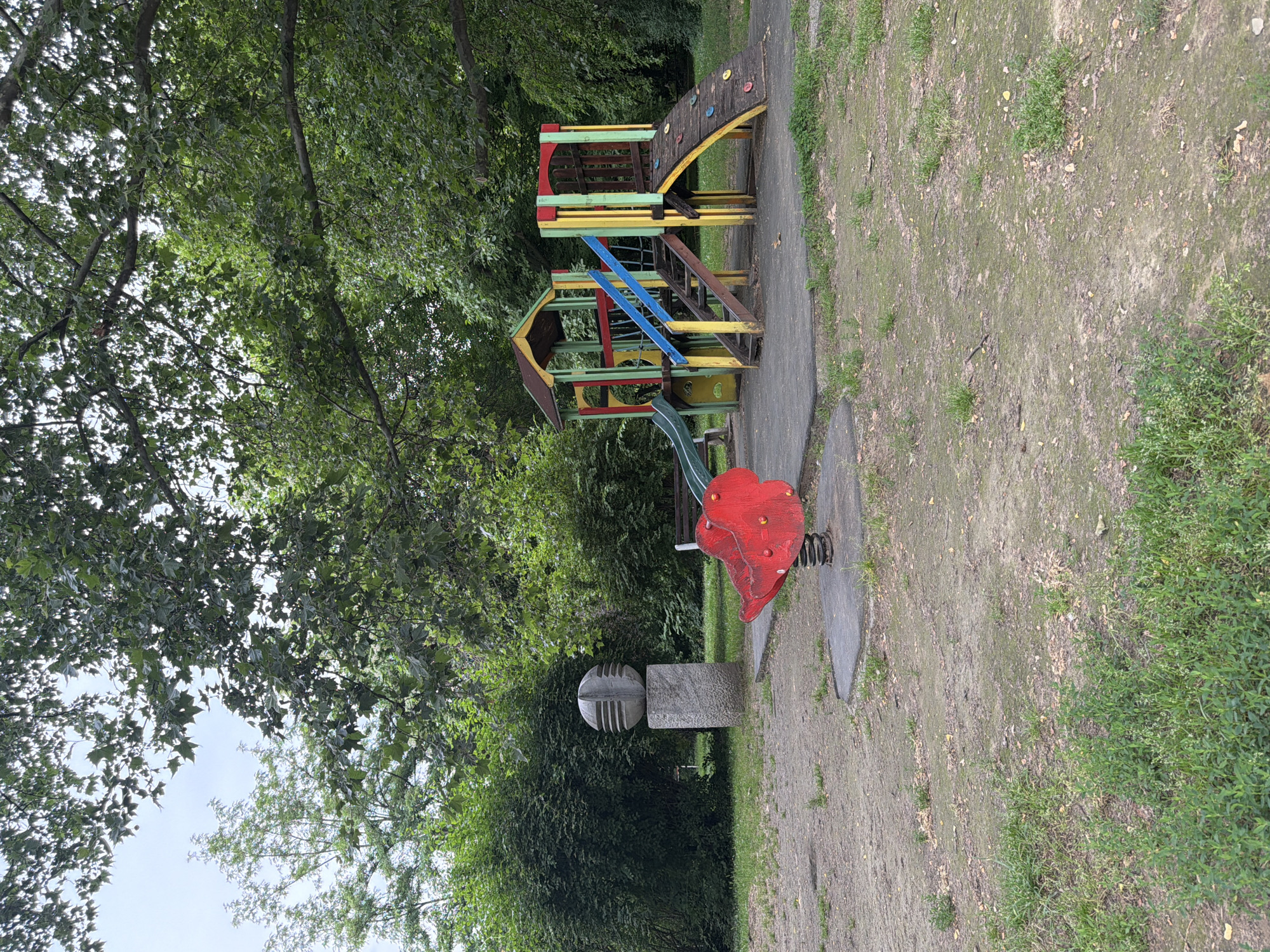 The image depicts an outdoor playground scene. In the foreground, a red, spring-mounted rocking toy shaped like an animal is visible. To the right, a multi-colored children's play structure features slides, climbing elements, stairs, and a small tower. Behind the play structure, trees and lush green bushes dominate the background, indicating a park or natural setting. To the left of the playground, a stone pedestal supports a spherical sculpture with horizontal grooves. The ground appears to be a mix of dirt, patchy grass, and fallen leaves. The sky is overcast, suggesting a cloudy day. The scene is located in a park in Pazardzhik, Bulgaria.