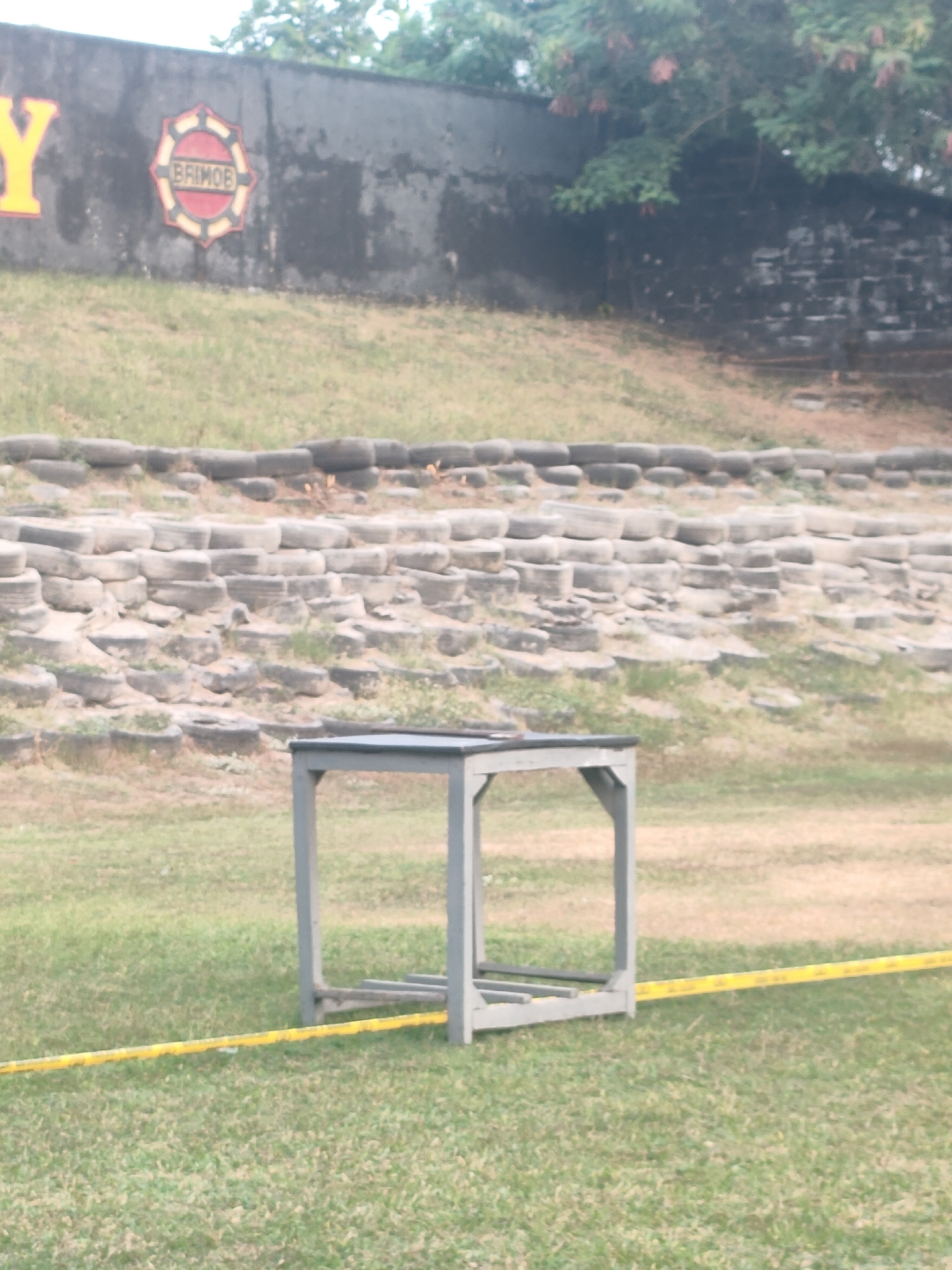 The image shows an outdoor training ground in Banguntapan, Indonesia. In the foreground, a grey plyometric box is placed on a grassy field. A yellow tape runs across the grass in front of the box. In the background, a wall with the text "BRIMOB" in yellow letters is visible. The wall is weathered and has a logo on it. Behind the wall, there are trees and vegetation. The ground slopes upwards from the wall, covered with grass and what appears to be a terraced structure made of tires. The overall scene suggests a training or obstacle course facility.