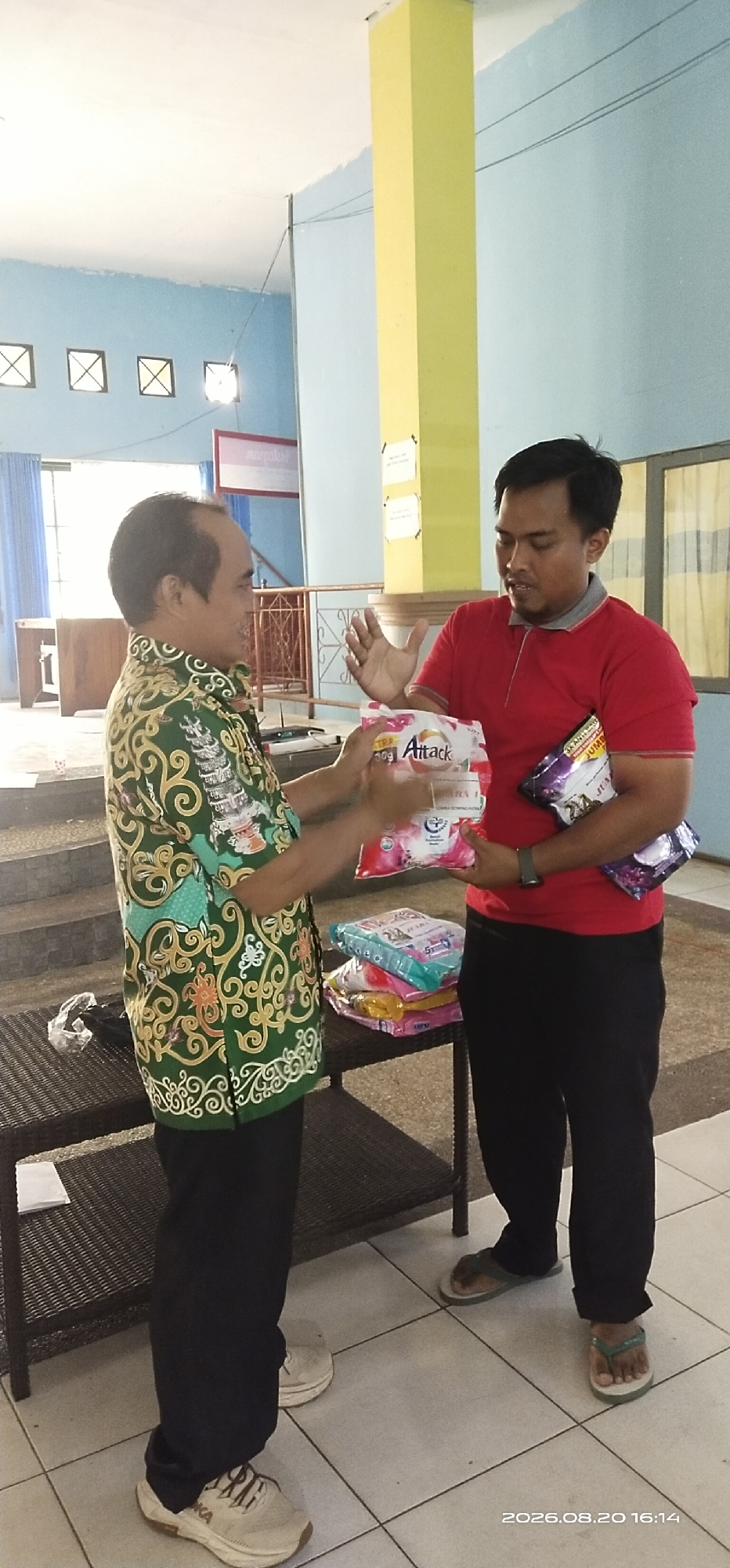 The image captures an indoor scene, likely a public building or community hall in Tamiang Layang, Indonesia, given the presence of a visible timestamp in the lower right corner that reads "2026.08.20 16:14". Two men are the central focus of the photograph, engaged in an exchange.
The man on the left, wearing a green batik shirt with intricate gold and white patterns and black pants, is facing the other man and gesturing with his hands. He appears to be in mid-conversation or handing something over. He is wearing white athletic shoes.
The man on the right is dressed in a red polo shirt with a grey collar and black pants. He is holding several packages of what appear to be cleaning or laundry products. One package in his right hand is clearly labeled "Attack". His left hand is raised, palm facing outwards, as if in explanation or emphasis. He is wearing flip-flops.
In the background, there is a yellow pillar and a light blue wall. The floor is tiled with light-colored squares. A dark brown woven shelf unit is positioned between the two men, holding more stacked packages of similar products. In the distance, architectural features like windows and a railing are visible, suggesting the location is a well-lit indoor space, possibly with an elevated section or stairs. The overall impression is that of a community event or distribution of goods.
