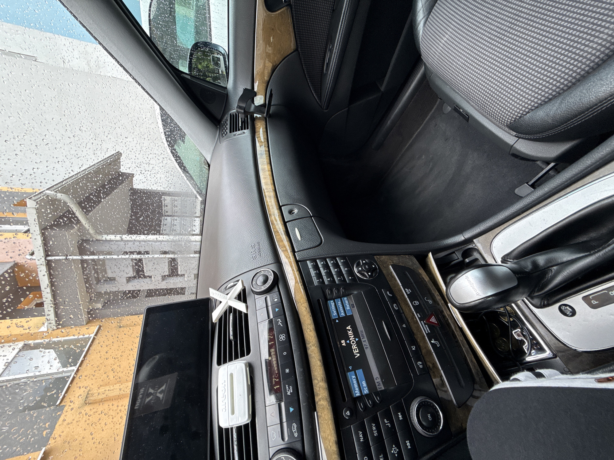 The image displays the interior of a Mercedes-Benz E-Class (W211, identified by the "Avantgarde" inscription on the gear selector and dashboard design) in Stara Zagora, Bulgaria. The perspective is from the driver's side, looking across the dashboard towards the passenger side and out through the windshield.
The windshield is heavily covered in raindrops, indicating active precipitation. Through the wet glass, several buildings are visible: an orange-yellow multi-story building on the left with prominent windows, and a lighter-colored building in the center-right featuring a rain gutter and a decorative or structural element. A portion of a white vehicle is faintly visible through the passenger window area.
Inside the car, the dashboard is prominent. An aftermarket tablet-style display screen is mounted on top of the central console, reflecting an "X" logo. Below this, an "Areon" branded air freshener and a separate white "X"-shaped decorative item are clipped to the central air vents. The car's climate control unit displays "17°C" on both sides. The integrated audio system's screen shows "VERONIKA" tuned to "97.4 MHz FM 5". A polished, light-brown wood trim panel extends across the dashboard and into the passenger door. A black phone mount is attached to the dashboard above the passenger glove compartment.
The center console features a silver-topped gear selector labeled "Avantgarde," currently in the "P" (Park) position, with a "W/S" mode switch visible. An open storage compartment next to the shifter contains several coins. The passenger seat is upholstered with a distinctive grey patterned fabric on its central sections, bordered by black leather. No people are visible within the frame.