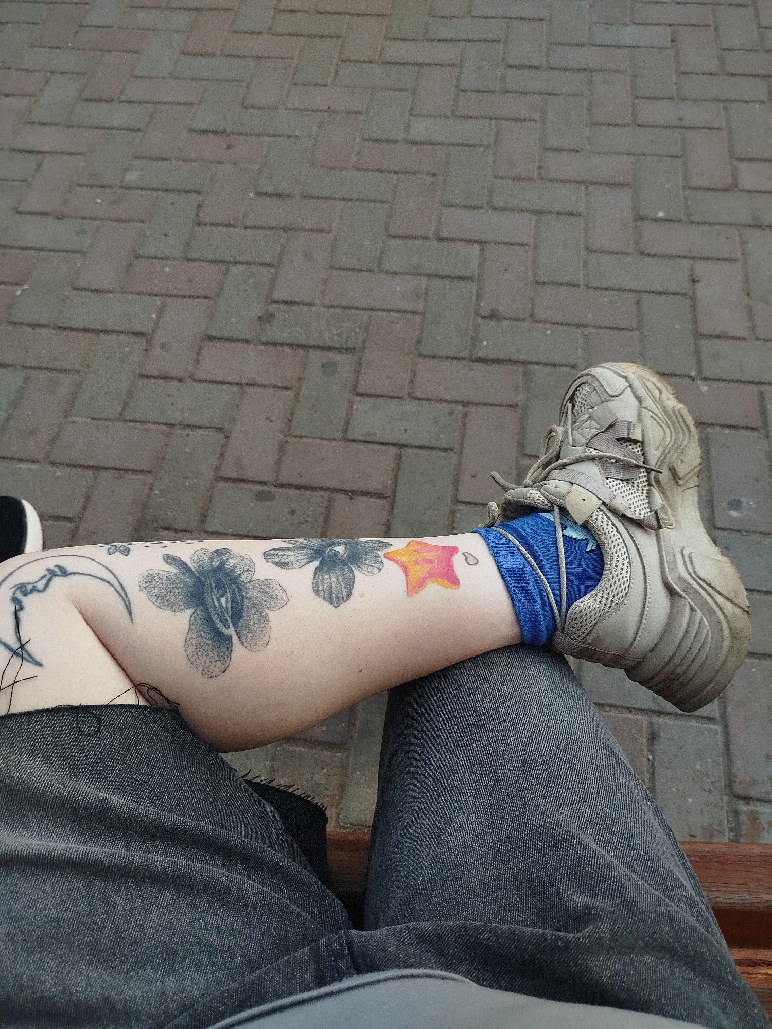 A first-person perspective image shows a person sitting on a bench, with their legs extended. The background consists of a brick-paved ground. On the person's left leg, which is bent at the knee, there are several tattoos, including a crescent moon with a face, two flowers, and a colorful star. The person is wearing dark denim jeans and blue socks. A beige sneaker with laces is visible on their right foot. The scene appears to be outdoors, possibly in a park or public space.