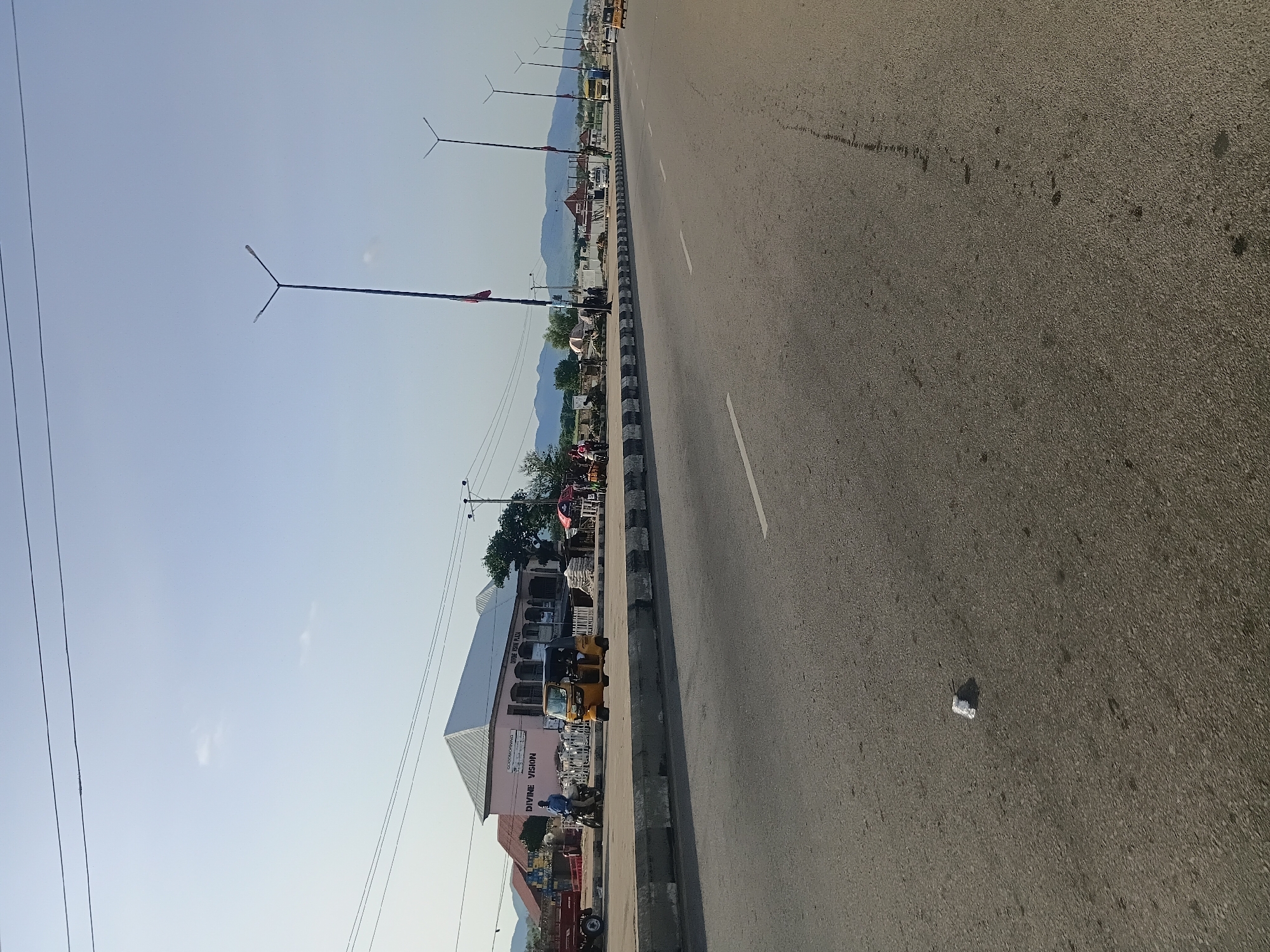 The image captures a wide asphalt road in Jalingo, Nigeria, likely during daylight hours with a clear sky. A concrete median with black and white reflective markings separates the lanes. On the side of the road, a pink building with a silver, sloped roof is visible, bearing signs that read "DIVINE VISION" and "DIVINE VISION PLAZA." In front of this building, several people are present. A man in a blue shirt is on a motorcycle, and a yellow auto-rickshaw (Keke Napep) is parked nearby. Numerous sacks of what appear to be cement or building materials are stacked along the roadside. Streetlights with multiple lamps are erected along the road's edge, some adorned with red banners. In the background, distant mountains frame the scene, and various vehicles, including a yellow truck, are seen on the road further down. The foreground of the road shows some debris, including a crumpled white item. The overall environment suggests a bustling commercial area within the city.