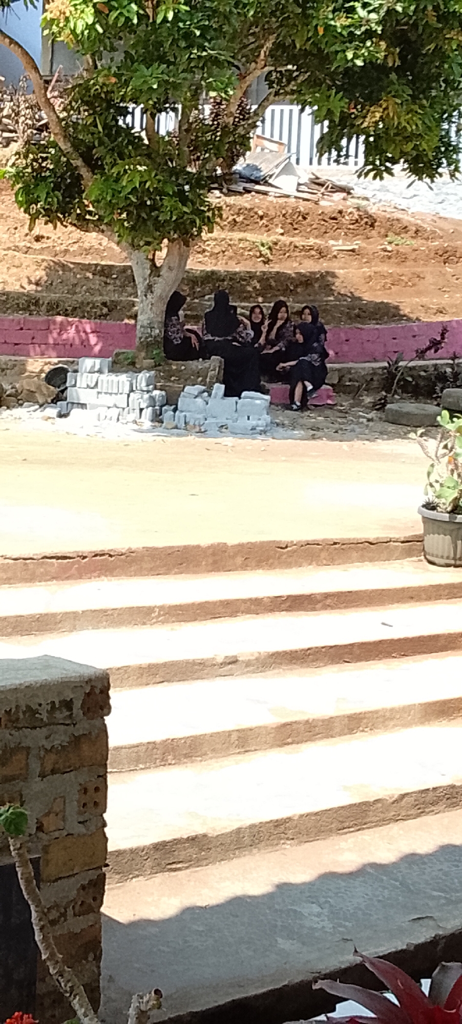 The image depicts a group of five young women dressed in dark clothing, possibly hijabs, gathered on a raised concrete and pink-bricked seating area beneath a tree. They are positioned on a tiered, outdoor space with steps leading down to a sandy or dusty ground. In the foreground, more steps are visible, constructed from concrete and brick. A potted plant with green leaves is situated on the right side of the frame. In the background, the scene suggests an outdoor environment with a dirt embankment, some scattered building materials, and a fence with vertical white stripes. The lighting indicates a sunny day, casting shadows across the steps. The location is identified as Tanjung Setia, Indonesia, suggesting a tropical or sub-tropical climate.