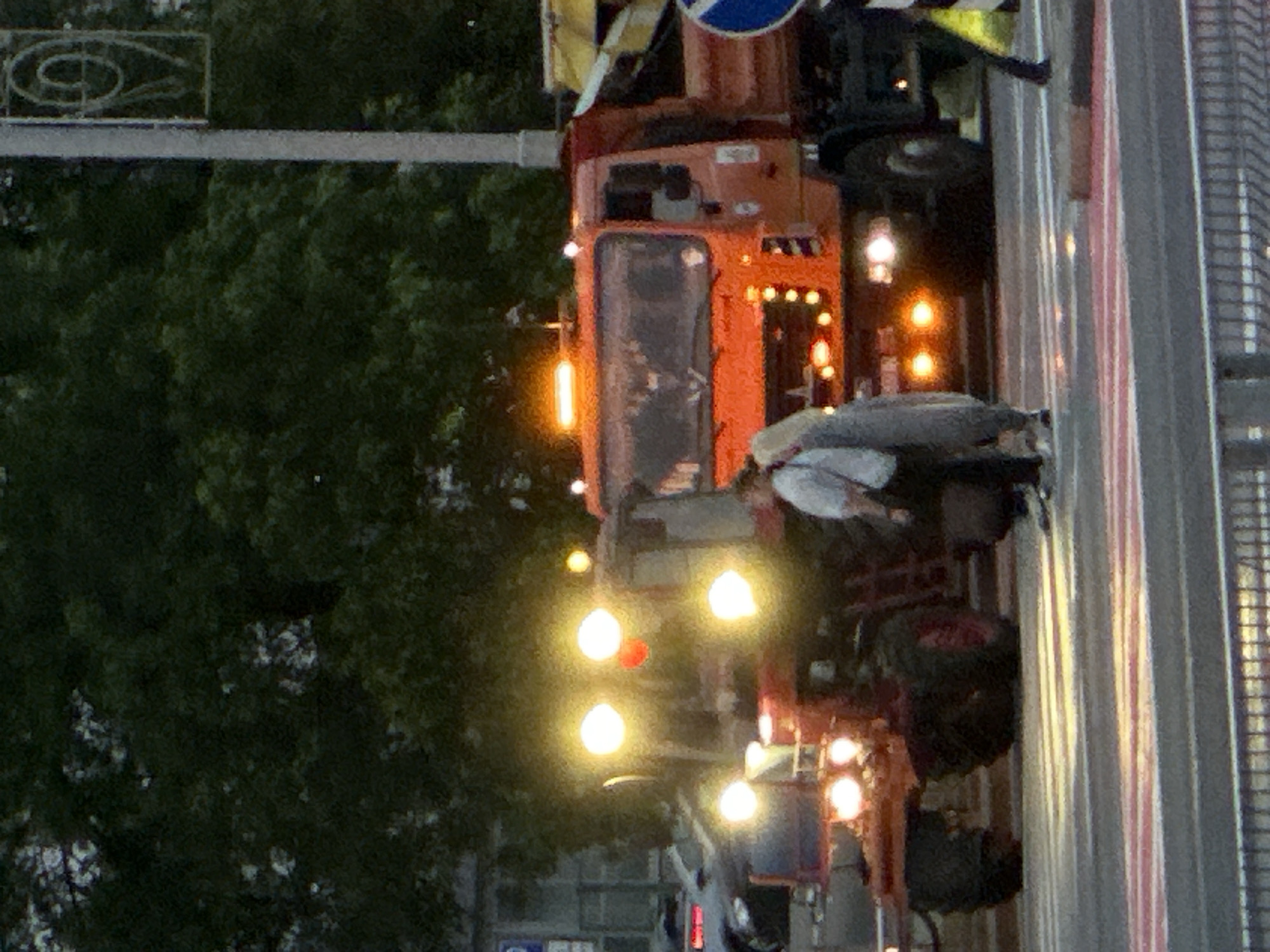 The image captures an urban road scene in low light, likely dusk or dawn, featuring two heavy vehicles and two individuals.
On the left, a red tractor, identifiable as consistent with a Belarus (MTZ) model, is stationary. Its front is illuminated by four bright, circular yellow/white headlights and additional auxiliary lights. To the right of the tractor, an orange dump truck, consistent with a MAZ or KAMAZ model, is also stationary. It features an illuminated amber warning light bar on its roof, main headlights, and several smaller amber lights on its front grille. A circular blue traffic sign is partially visible to the right of the truck.
Two individuals are positioned between the tractor and the truck, standing on a pedestrian crossing. The person on the left wears a white shirt and dark trousers, facing towards the right. The second person, on the right, has long blonde or light-colored hair and is dressed in a light grey or beige outfit, facing the first individual. They appear to be in conversation or interacting. A third person's lower leg and foot are visible on the extreme right of the frame, near the blue traffic sign.
The road surface in the foreground displays distinct red and white striped markings, indicating a pedestrian crosswalk, a common feature in Minsk, Belarus. The ground also shows reflections from the vehicles' lights, suggesting a wet or reflective surface. In the background, dense green foliage from trees is visible, along with a grey street light pole featuring an ornamental, scrolled metal top. A faded blue 'P' sign (parking) is faintly discernible in the far left background. The overall scene indicates a temporary stop or interaction on an urban street in the city of Minsk, Belarus.