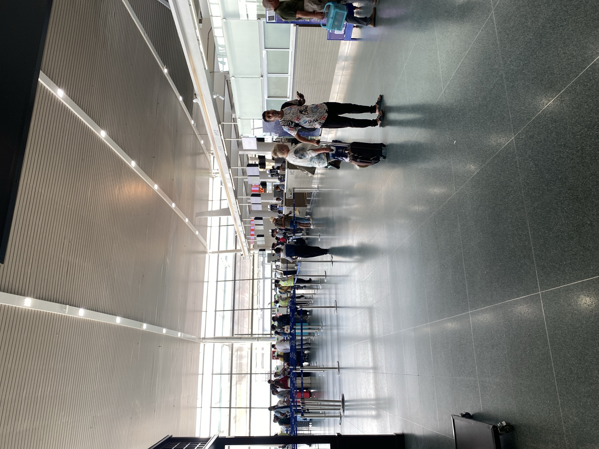 The image captures a bustling, well-lit airport terminal, likely a check-in or security area, located in Mai Khao, Thailand. The scene is characterized by its modern, spacious design, featuring a high ceiling with linear lighting fixtures and light-colored, possibly wooden, slatted panels. Large windows run along one side of the terminal, allowing ample natural light to illuminate the space, suggesting it is daytime. The floor is a dark, speckled material, possibly granite or polished concrete, with visible tile lines.
Numerous people are present, primarily standing in queues that extend down the length of the hall. These queues are defined by metal stanchions. Many individuals have luggage, including various rolling suitcases. In the mid-ground, a young man, wearing a distinct patterned short-sleeved shirt, dark trousers, and sandals, faces the camera with one arm raised, possibly posing. Text on his shirt is partially visible but indistinct. Near him, an older woman with light-colored hair is seen with a dark rolling suitcase. Further down the hall, more passengers queue, some appearing to be at check-in desks.
Notable details include the sleek architecture and the orderly lines of people. On a structural element or pillar, the labels "H11" and "H12" are faintly visible, likely indicating gate numbers or check-in counters. A blue plastic basket is partially visible near the top right, and a black monitor screen can be seen in the bottom right corner. The overall impression is one of organized transit activity within a clean and contemporary airport environment.