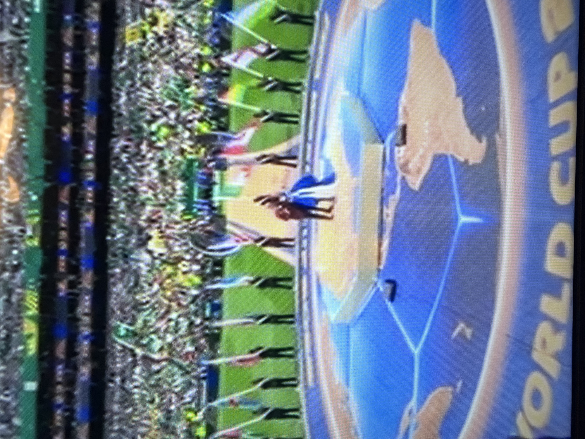 The image depicts a large stadium filled with spectators. In the foreground, a circular platform with a world map design is visible on the ground, with the words "WORLD CUP" emblazoned on it. Several people are standing on this platform, some holding flags and others in the process of what appears to be a ceremony. Two individuals are at the center of the platform, one in a blue dress and the other in red. A line of individuals, each holding a flag, are positioned along the edge of the platform. The stadium seating is filled with a crowd, and flags of various colors are scattered throughout the stands. The lighting suggests an outdoor event, likely during the day.