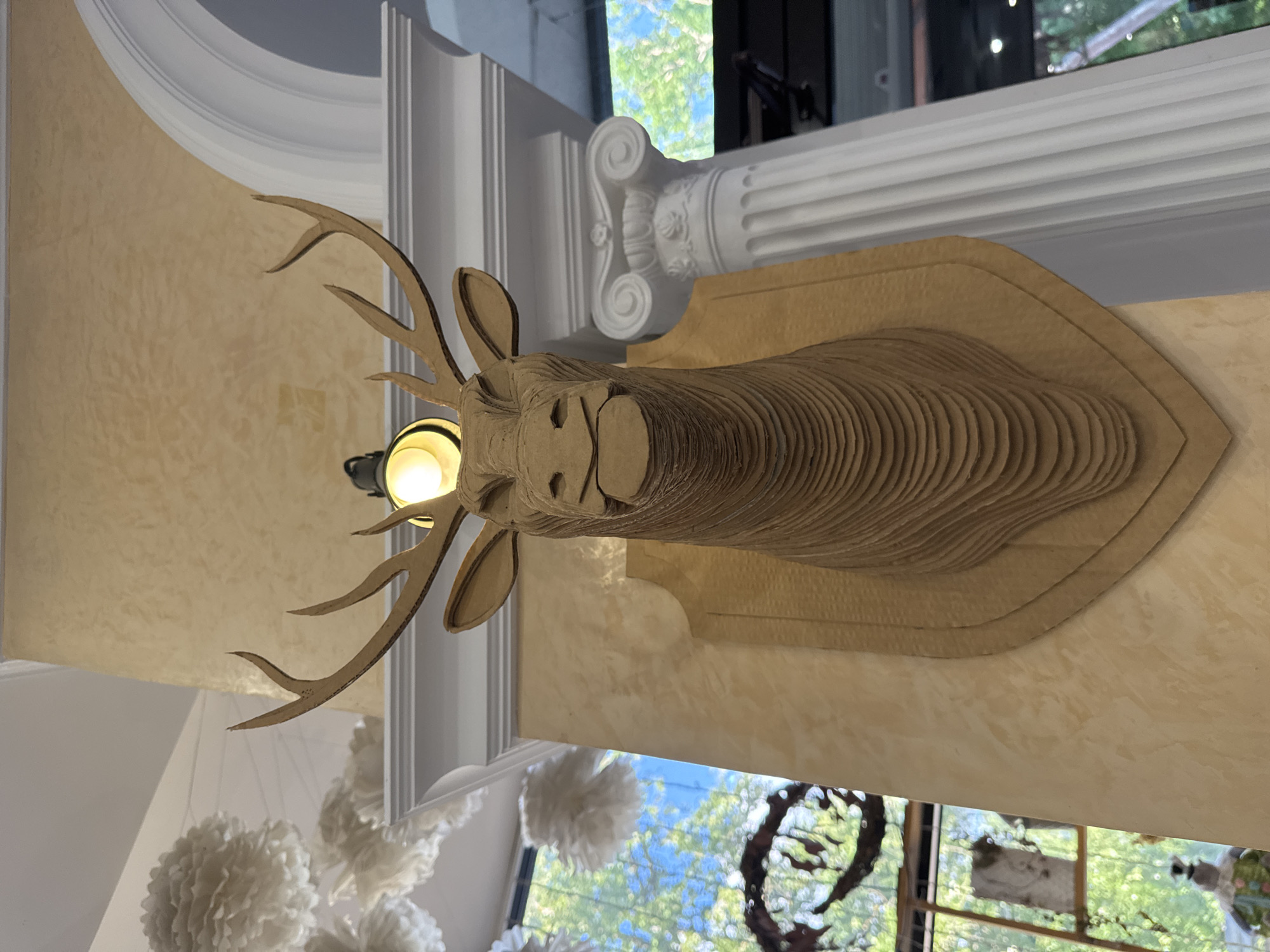 The image displays a faux taxidermy deer head crafted from layered cardboard, mounted on a shield-shaped plaque. The deer head has large, stylized antlers and a simplified facial structure. It is affixed to a light tan, textured wall. Above the deer head, a vintage-style lamp with a glowing bulb is visible. To the left of the deer head, decorative white paper pom-poms hang from the ceiling. To the right, a white Corinthian column stands prominently. In the background, through a window, lush green trees and blue sky are seen, suggesting an outdoor or garden view. The scene appears to be an indoor decorative display, possibly in a cafe, restaurant, or shop. The specific location within Saint Petersburg, Russia, is not discernible from the image itself.