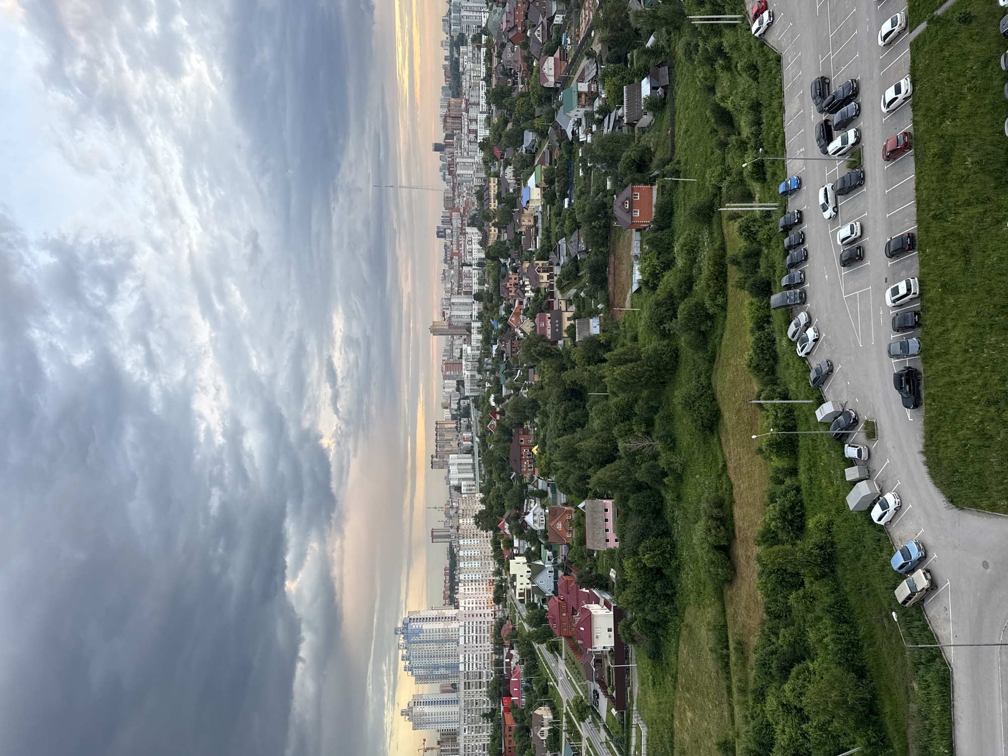 This is an aerial, wide-angle, vertical shot of a mixed urban and suburban landscape under a cloudy sky, likely taken at dusk or dawn. The scene is dominated by a sprawling residential area with a mix of high-rise apartment buildings in the distance and smaller, individual houses with red-tiled roofs closer to the foreground. Dense green foliage, including trees and grassy fields, separates the housing developments. A parking lot filled with various cars, including sedans, SUVs, and vans, is visible in the lower portion of the image, adjacent to a grassy area and a road. The sky is characterized by dramatic, dark grey clouds with some lighter patches suggesting diffused sunlight. The overall impression is one of a developing urban edge, with modern high-rise buildings encroaching on a more traditional suburban or village setting. Based on the architectural style and the presence of many trees, this could be a scene from the outskirts of Perm, Russia, possibly in a district undergoing development.