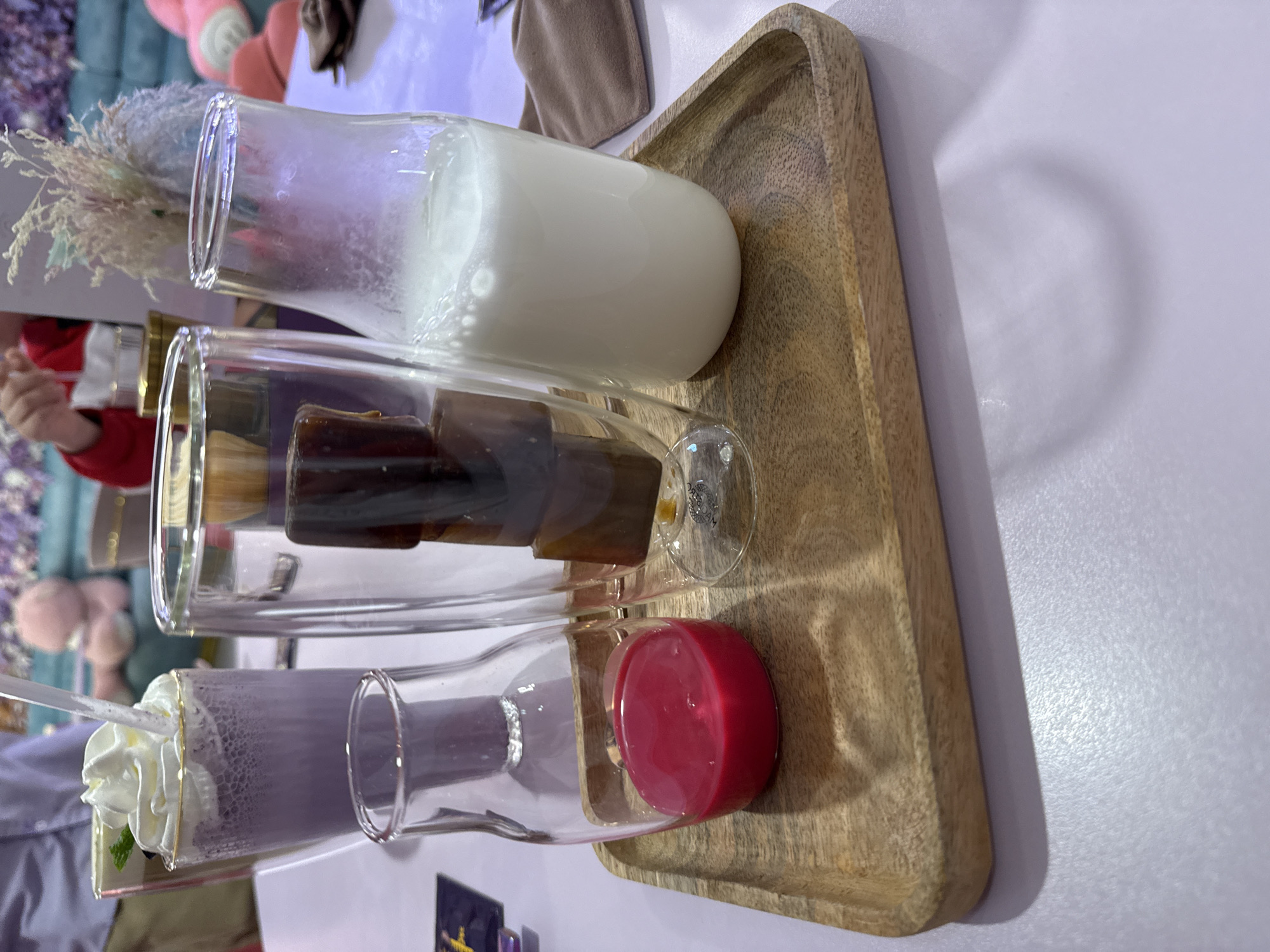 On a wooden tray are three clear glass vessels, presumably containing beverages. The top vessel holds a white, opaque liquid. The middle vessel is filled with ice cubes. The bottom vessel contains a purple liquid with a dollop of whipped cream on top. The scene appears to be indoors with additional decorations in the background. There is no indication of this image being in Vladivostok, Russia.