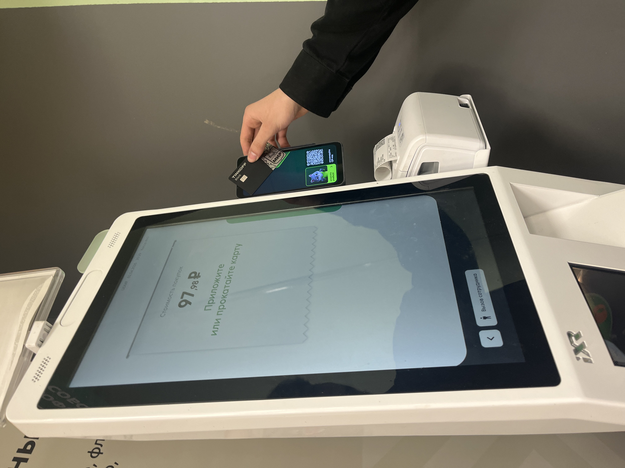 A photograph shows a hand holding a black Tinkoff bank card and a phone over a self-service kiosk screen with the message in Russian, “Attach or swipe card” and the amount 97,98 rubles. A receipt printer is next to the phone. The brand iXE! is visible on the kiosk. The kiosk screen also indicates the possibility of calling an employee. The setting is likely in a store in Russia, as the text on the screen is in Russian and the card is from Tinkoff bank. The specific location within the city of Chaikovsky, Russia, cannot be determined from the image alone.