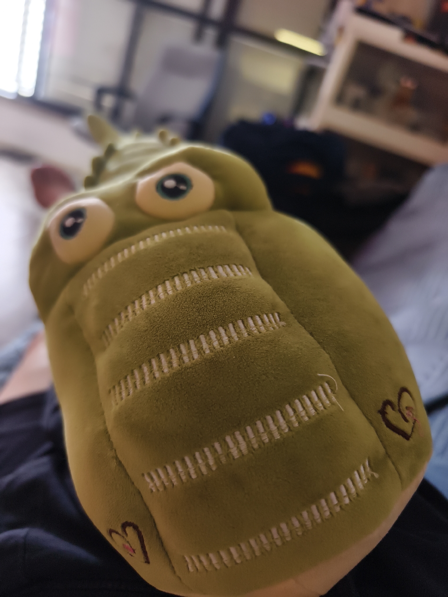 The image is a close-up shot of a green stuffed alligator toy. The toy has large, cartoonish eyes and white, stitched lines across its body, with small heart symbols embroidered on its sides. The background is blurred but appears to be an interior setting, possibly a home. It is unclear if the image was taken in Petah Tikva, Israel. The toy is resting on a person's lap.