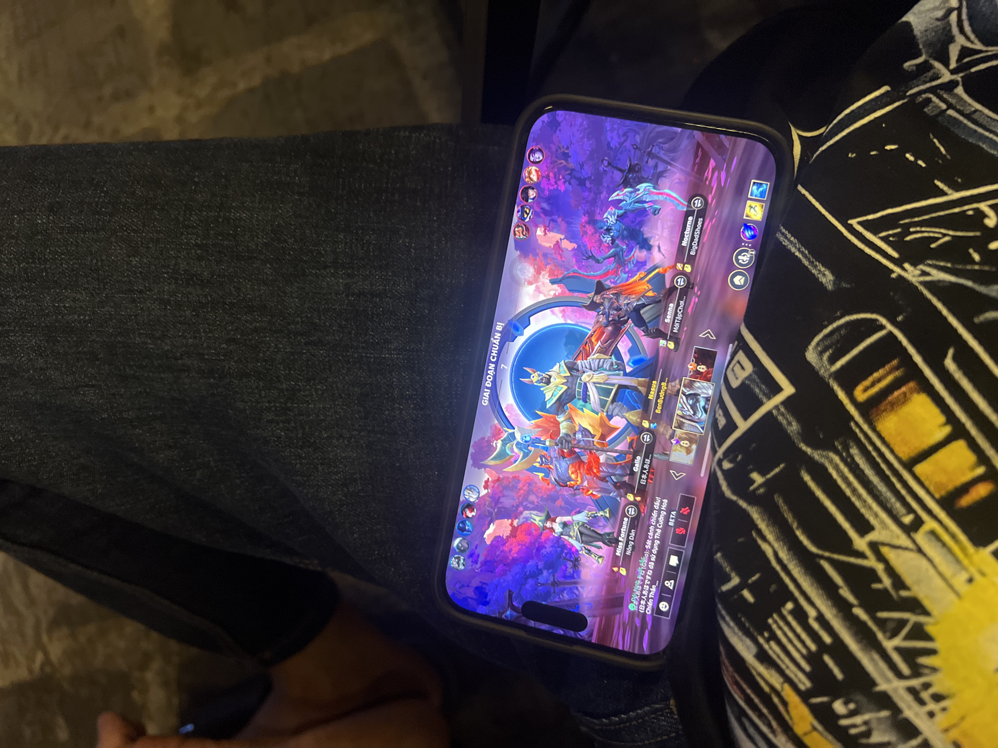 A person is holding a mobile phone, displaying the Teamfight Tactics mobile game. The phone is positioned on the person's lap, which is covered in denim pants and a patterned fabric. The game screen shows a dynamic scene with various characters and game elements, including character names like "Miss Fortune" and "Nocturne." Text in Vietnamese is also visible. The location appears to be indoors, given the lighting and proximity to the person. There is no explicit information in the image regarding Đà Nẵng, Vietnam.