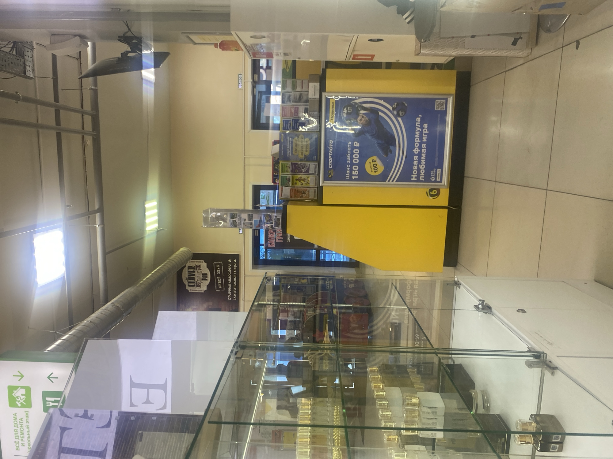 This image depicts the interior of a commercial building, likely a store or shopping center, in Chaikovsky, Russia. The scene is dominated by store fixtures, including a long, yellow counter or display stand on the right, and multiple glass display cases in the foreground on the left. The floor is tiled, and artificial lighting from overhead fixtures illuminates the space. A large, reflective air duct runs across the ceiling.
No people are visible, but an advertisement on the yellow stand features an image of a man. The activity appears to be the display of goods or services. The glass cases in the foreground contain numerous small, uniform bottles or containers, possibly cosmetics, perfumes, or similar products.
Visible text provides several clues about the location and offerings:
* A prominent sign reads "ВСЁ ДЛЯ ДОМА И РЕМОНТА (цокольный этаж)," indicating a home improvement section on the basement floor.
* Further into the background, a "CLOVER BAR" sign is visible, along with phrases like "ЖИВОЙ ЗВУК" (LIVE SOUND), "ОТЛИЧНАЯ АТМОСФЕРА" (GREAT ATMOSPHERE), and "ЗАЖИГАТЕЛЬНЫЕ ТАНЦЫ" (ENERGETIC DANCES), suggesting entertainment.
* The yellow stand features a large advertisement for "СПОРТЛОТО" (SPORTLOTO), a lottery, with text promising "Шанс забрать 150 000 Р" (Chance to win 150,000 Rubles) for "Всего за 100 Р" (Only for 100 Rubles). It also highlights "Новинка" (New) and "Новая формула, любимая игра" (New formula, favorite game).
* An "выход" (exit) sign is visible above a doorway in the background.
* Other partially visible signs include "БАНКР ГРАХ ЗАКОНН" (possibly related to legal/financial services), "БЫТ" (LIFESTYLE), "PIKOU," and "SOIN RA."
The time of day is not discernible due to the indoor setting and artificial lighting. The overall impression is that of a bustling commercial environment offering a variety of products and services.