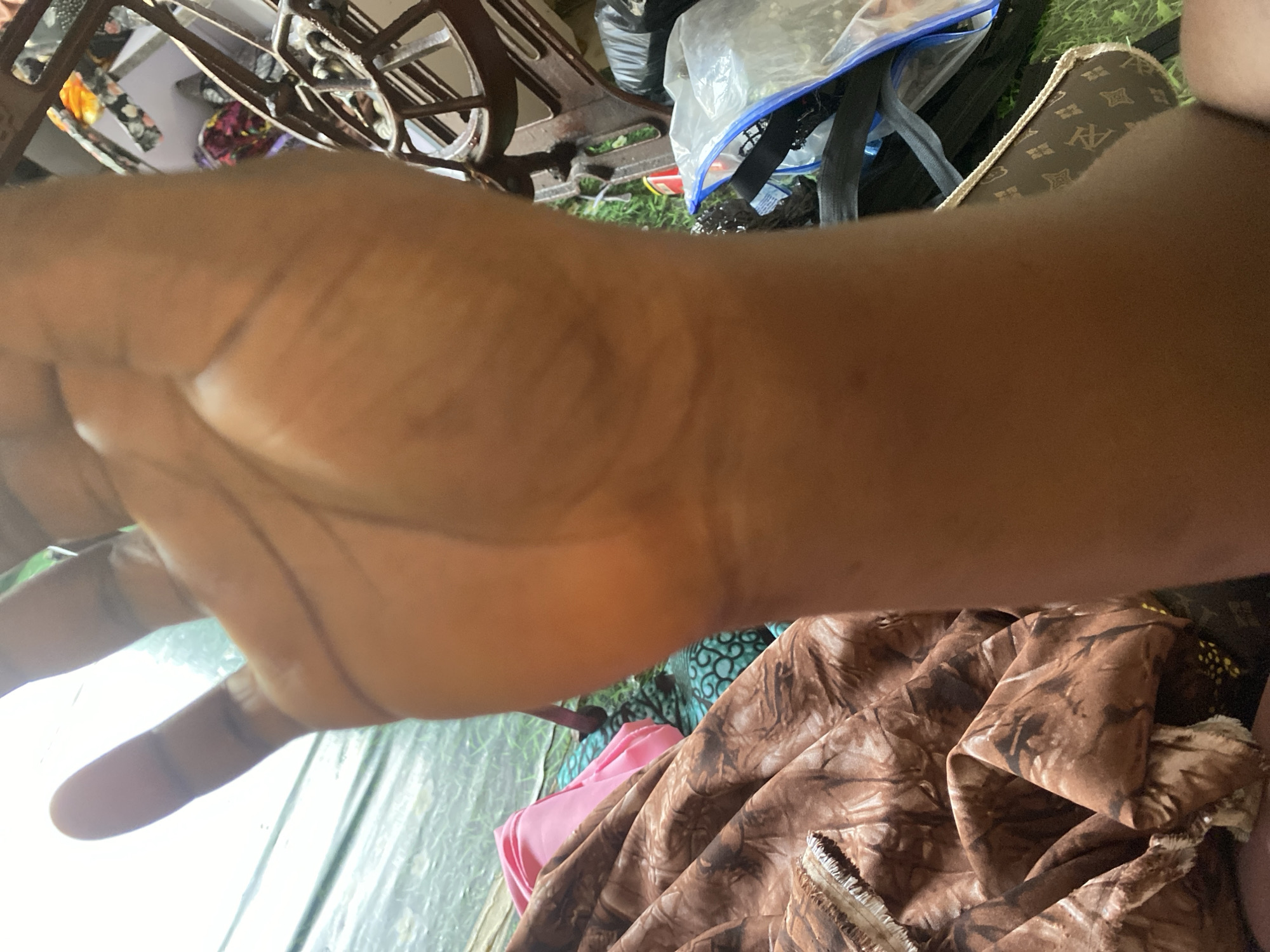 A close-up, vertical, first-person perspective shot shows a dark-skinned hand held up, with the palm facing the camera and the thumb extended upward. The hand is in focus, with visible lines and creases on the palm. The background is blurred but suggests an indoor setting with various objects. To the left of the hand, fabric with a brown and white abstract pattern is visible, along with a folded piece of pink fabric. Further to the right, a vintage sewing machine and several plastic bags are present. The floor appears to be covered in green material, possibly grass or a rug. The lighting is bright, casting shadows and highlighting the skin texture. The image is taken in Jauro Yinu, Nigeria.