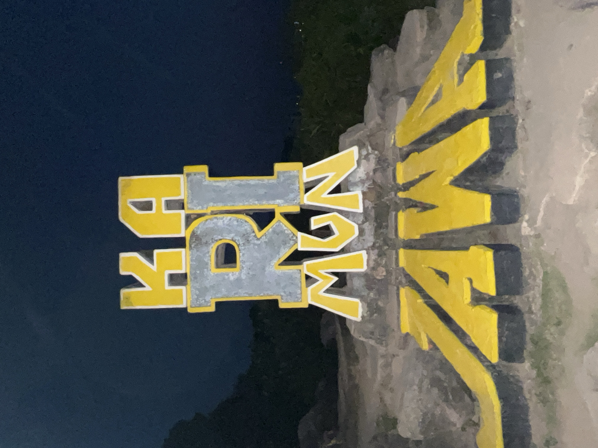 The image displays a large, prominent sign with the words "KARIMUN JAWA" in bold, three-dimensional lettering. The letters are primarily yellow with white and gray accents, and they are mounted on a rough, stone-like base. The sign appears to be a landmark or welcome sign for the location of Karimunjawa, an archipelago in Indonesia known for its marine national park. The background is dark, suggesting it is nighttime or dusk, with the sky a deep blue-gray. There are subtle indications of foliage and rocky terrain in the background, consistent with a natural or semi-natural setting. No people are visible in the image.