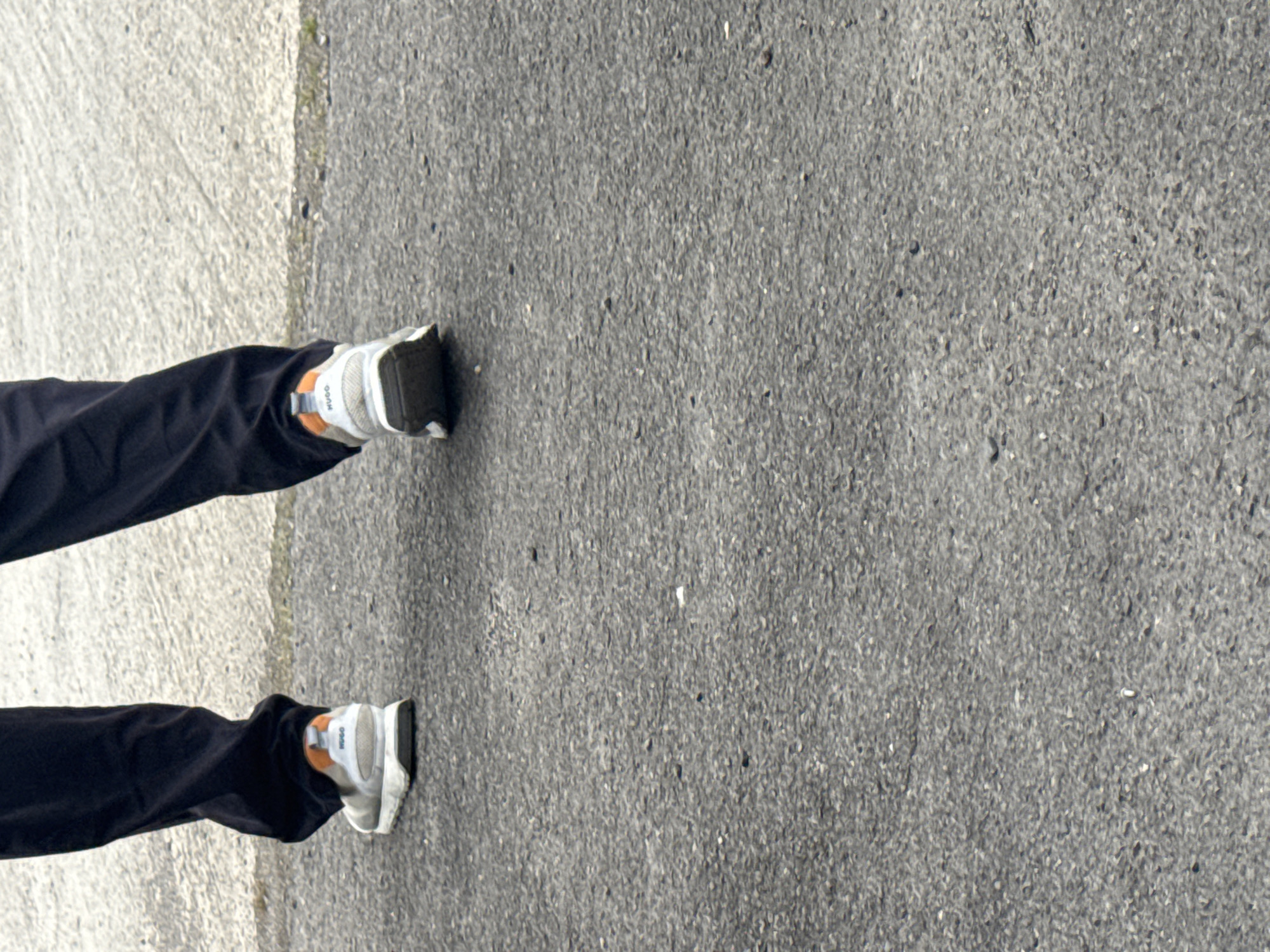 The image shows a close-up view of a person's legs and feet walking on an asphalt surface. The person is wearing dark trousers and grey sneakers with orange accents on the heels. The sneakers appear to be branded with the word "HUGO". The background is a textured, light-colored surface, possibly concrete. The overall scene is generic and does not contain any specific landmarks or elements that would allow for precise identification of the location within Tewkesbury, United Kingdom. The image appears to be taken outdoors during the daytime.