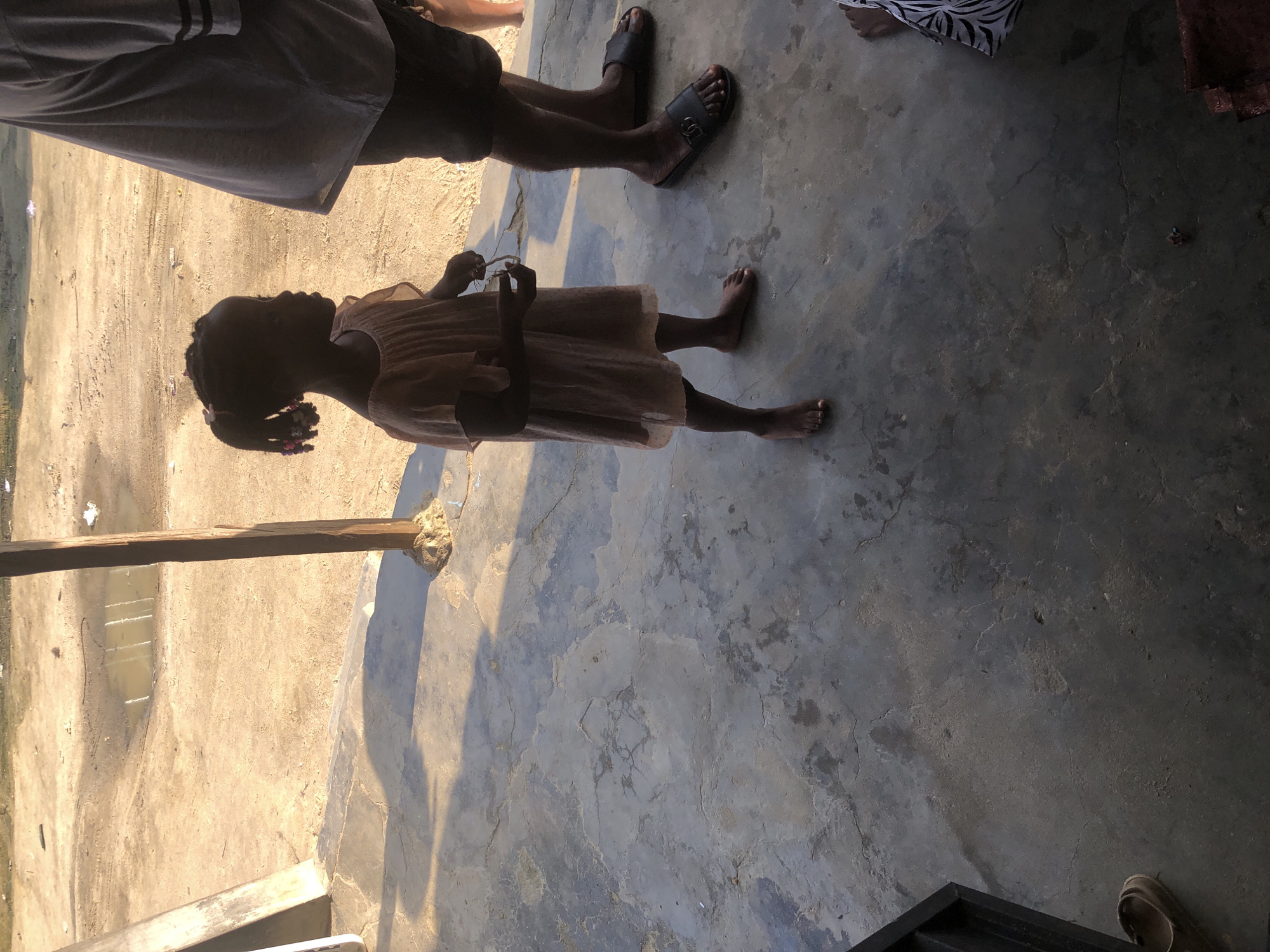 A young Black girl in a light-colored, gauzy dress stands on a cracked concrete surface in what appears to be Ado, Nigeria. Her hair is pulled into two pigtails with colorful beads. She is holding a small object in her right hand. To her right, a person's legs are visible; they are wearing dark shorts and black slides with a double G logo. In the background, a sandy area with a puddle of water is visible. A wooden pole supports an overhead structure, casting a long shadow across the concrete. The overall lighting suggests a sunny day.