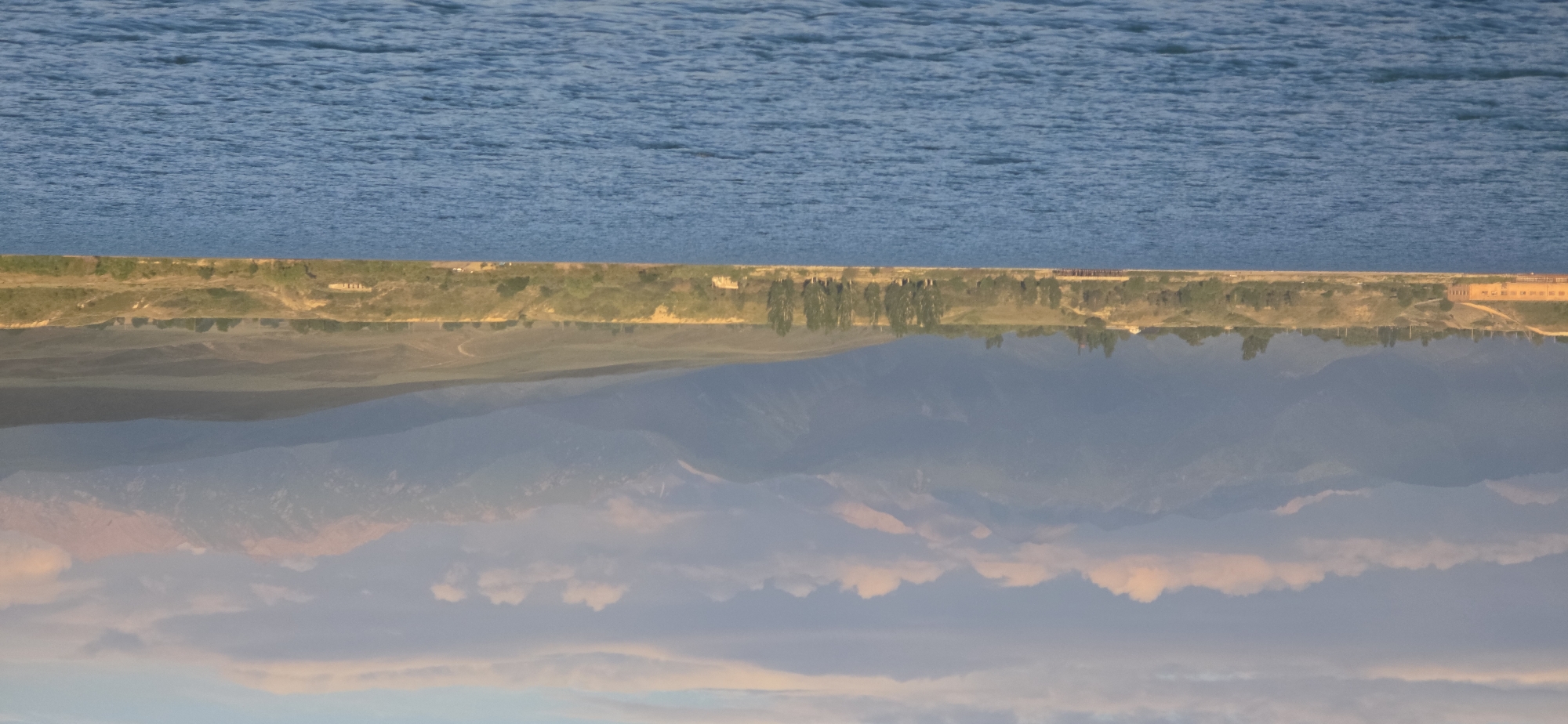 This image depicts a serene lake with a mountainous backdrop. In the foreground, the deep blue water of Issyk-Kul Lake is visible, with gentle ripples disturbing its surface. The middle ground features a shoreline lined with trees and vegetation, interspersed with some structures. On the left side of the shoreline, a long, low building with a light-colored facade is prominent. Further along the shore, a wooden pier extends into the water. Beyond the shoreline, rolling hills lead up to the imposing mountain range that dominates the background. The mountains are layered in shades of blue and grey, with some peaks catching the soft light of what appears to be late afternoon or early morning, casting pink hues on some of the higher elevations. The sky above is a soft pastel, with scattered clouds. The scene is tranquil and appears to be a natural landscape with some signs of human presence near the lake. This location is within or near the city of Pristan'-Przheval'sk, Kyrgyzstan, on the eastern shore of Issyk-Kul Lake.