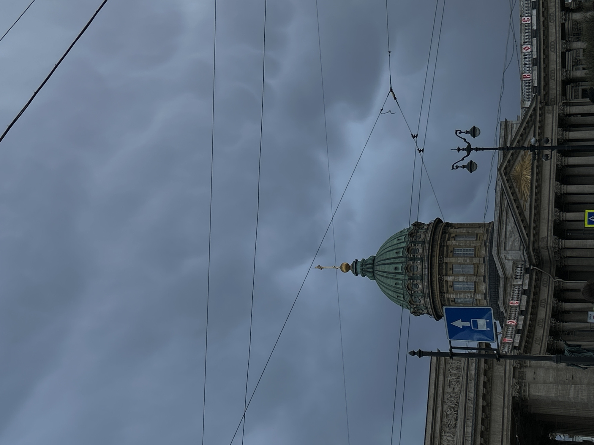 The image displays a partial view of the Kazan Cathedral in Saint Petersburg, Russia, under a heavily overcast sky. The cathedral's prominent green dome, adorned with a golden finial and a cross, rises above the building's classical facade, which features columns, ornate carvings, and windows.
Numerous dark overhead wires, characteristic of urban public transport infrastructure, crisscross the gloomy, cloud-filled sky. An ornate black street lamp with two lights is positioned in the mid-ground. Two distinct street signs are visible: a blue square sign with a white arrow pointing left and a bus icon, indicating a bus stop or lane; and a yellow square sign with a black pedestrian crossing symbol and two downward arrows.
On the upper part of the cathedral's facade, red square banners or signs with faint white symbols are discernible. Below these, illuminated red digits display "19" and "03," possibly indicating the time or temperature. The weather appears dull and cloudy, contributing to a muted color palette across the scene. No people or specific activities are clearly visible, focusing the image on the grand architecture and urban elements.