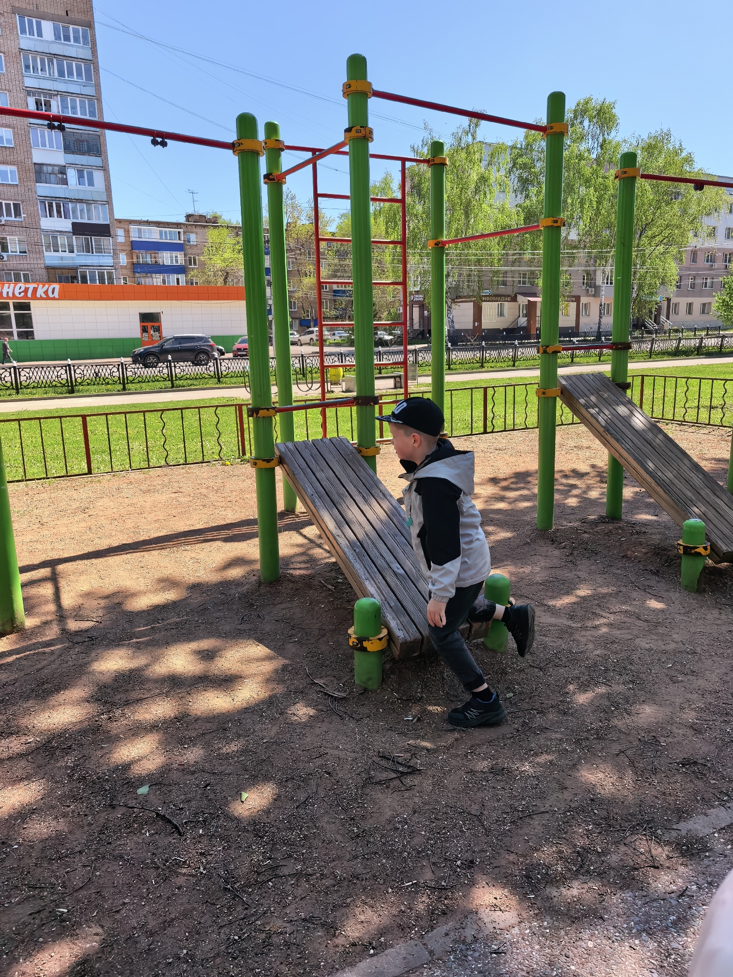 The image depicts a young boy at an outdoor exercise park in Sterlitamak, Russia. The boy is wearing a black cap, a light gray jacket with black sleeves, dark pants, and black shoes. He is standing on a wooden plank attached to green and yellow supports. The park features other exercise equipment, including monkey bars and inclined benches. The ground is dirt and mulch. In the background, there are apartment buildings, including one with an orange sign that reads "Herka". A green fence separates the park from the street, where cars are visible. The sky is bright blue, indicating a sunny day.