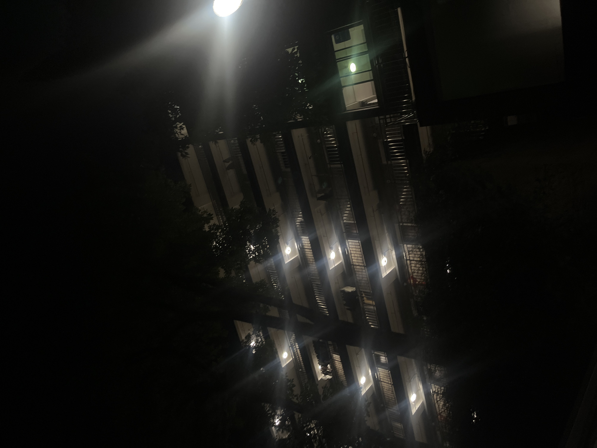 The image depicts a multi-story residential building at night in Yuzhong District, China, viewed from a low angle. The building features numerous balconies with metal railings across multiple floors. Many balconies are illuminated by overhead lights, casting visible light flares. On several balconies, items resembling clothes are hanging, suggesting laundry is drying. Dark tree foliage partially obscures the building on the left and center. A bright street light is prominently visible on the far right, emitting a strong light and lens flare effect, illuminating a portion of the upper right quadrant of the image. One window on the lower right section of the building is lit from within, revealing a green light fixture. The overall scene is dark, characteristic of nighttime, with artificial illumination highlighting parts of the building and the immediate surroundings. No individuals or specific actions are discernible beyond the hanging clothes.