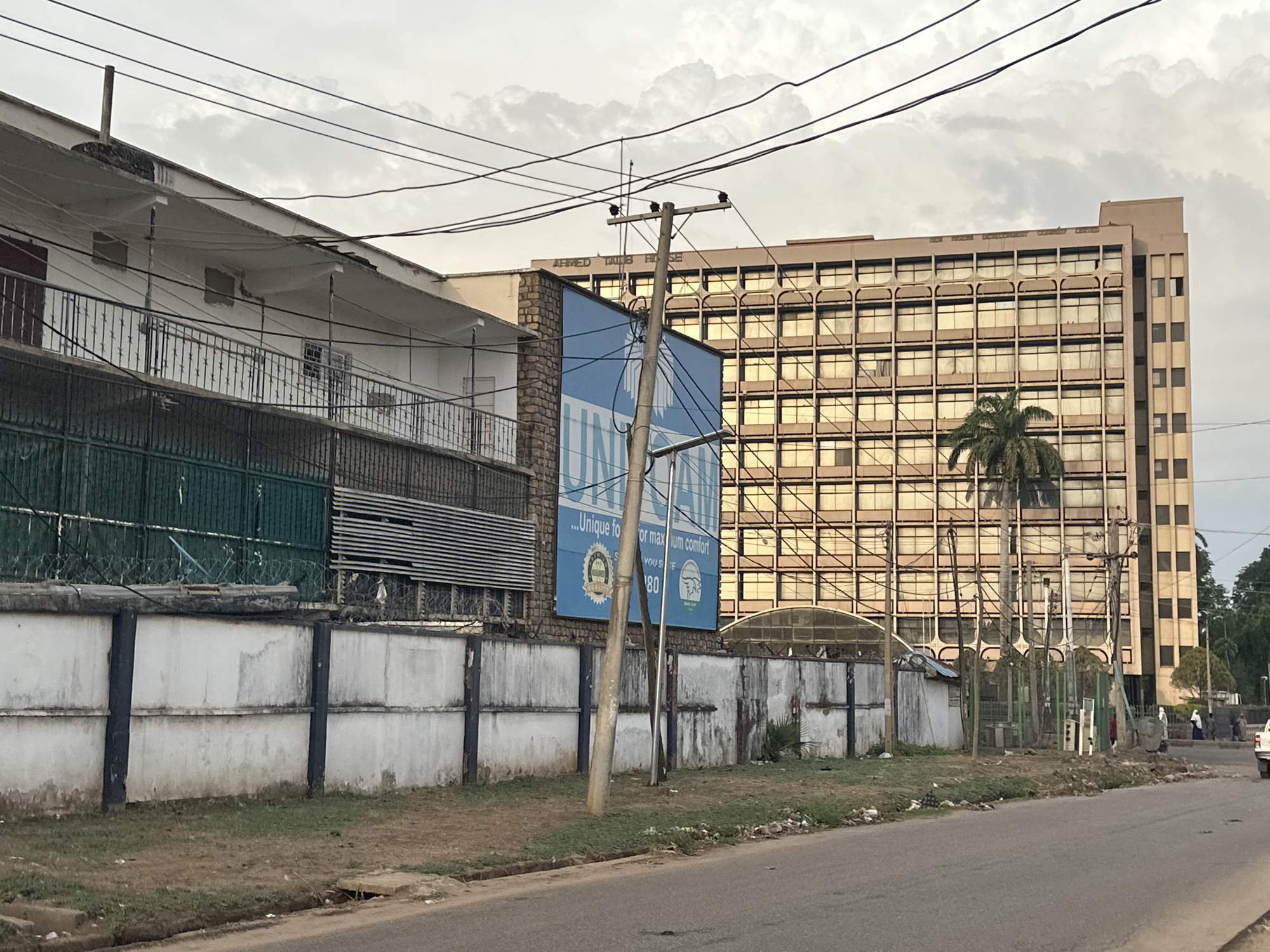 The image depicts a street scene in an urban location. A multi-story building, possibly an office complex, is on the right, with a grid of windows reflecting sunlight. A palm tree stands in front of the building. An older, two-story building is on the left, behind a concrete wall topped with fencing and barbed wire. A large advertisement is mounted on a pole in front of the wall. Overhead power lines crisscross the sky. The street appears to be paved but worn, with litter and debris along the curb. A few people and vehicles can be seen in the background. The sky is overcast with clouds.