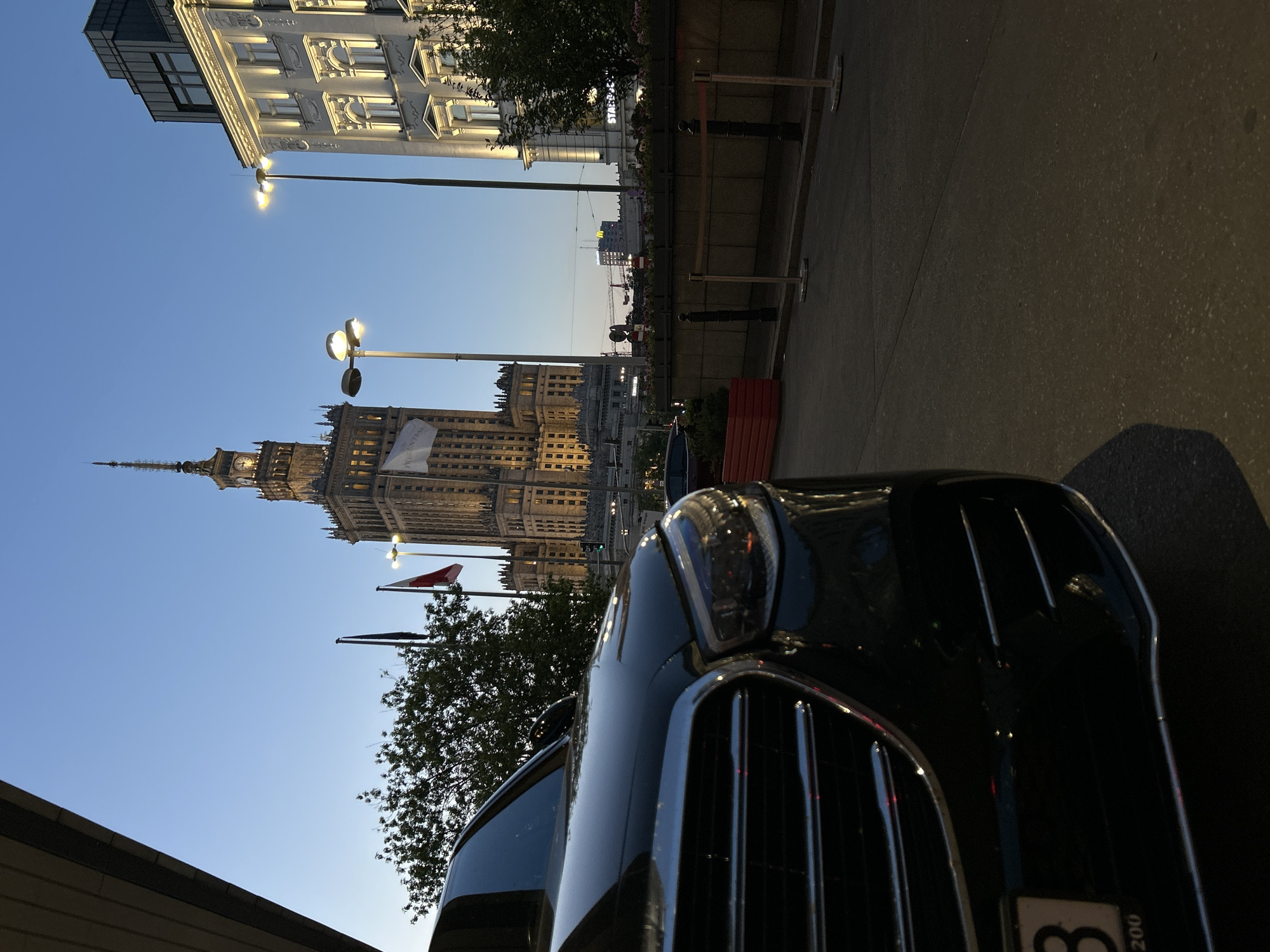 The image shows a low-angle shot of a black car parked on a street in Warsaw, Poland. In the background is the Palace of Culture and Science, a prominent landmark in Warsaw. The sky is light blue, suggesting it is either early morning or late evening. There are street lights and Polish flags visible near the Palace. The upper part of a building is seen at the top of the image.