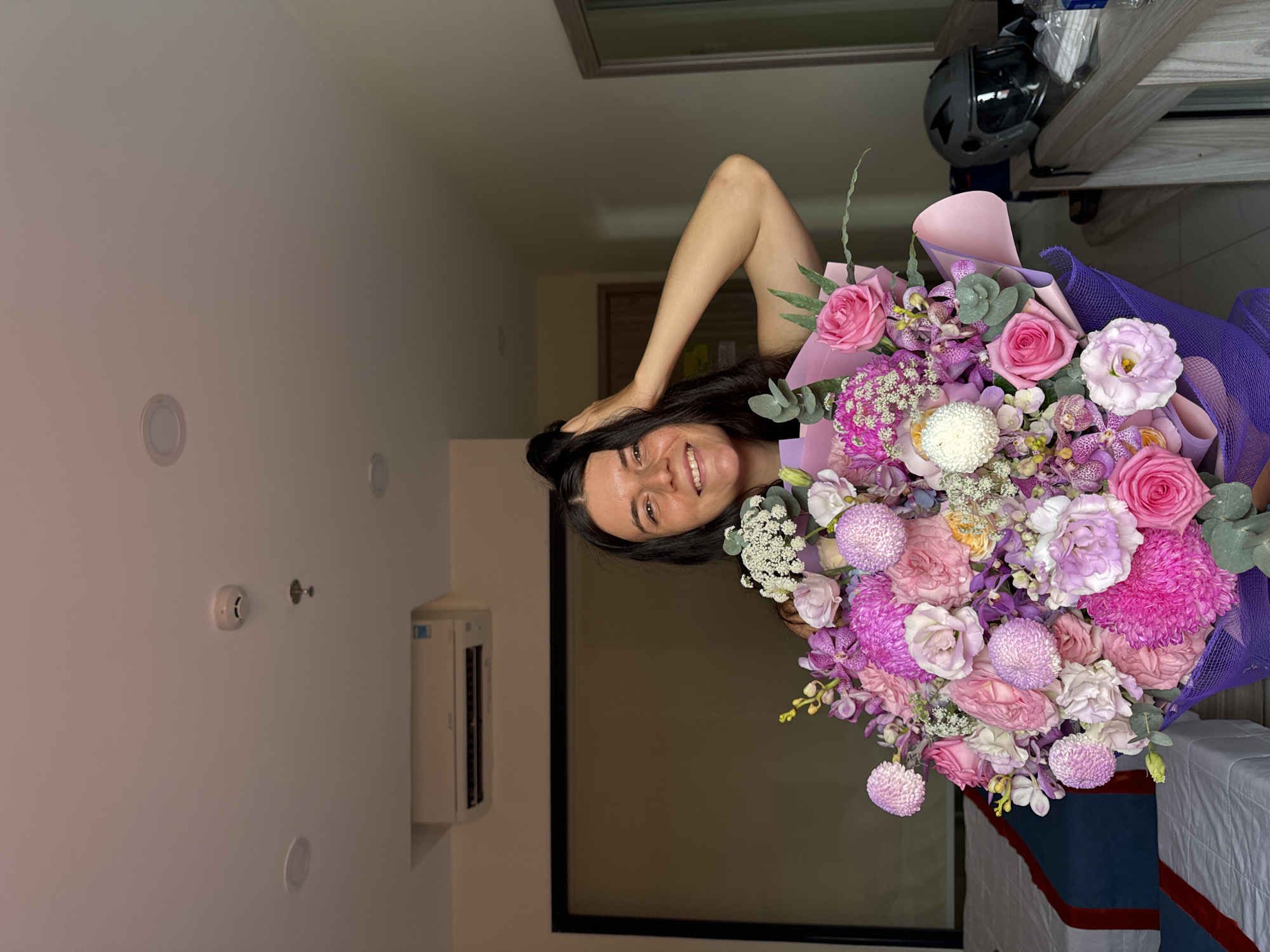 A young Caucasian woman with long, dark hair is holding a large bouquet of pink and purple flowers in a hotel room. She is smiling and touching her hair with her right hand, which is raised above her head. The bouquet is composed of various types of flowers, including roses, chrysanthemums, and orchids. The room has a neutral color palette with a white ceiling, light-colored walls, and a dark carpet. An air conditioning unit is mounted on the wall above a window with black framing. A bed with white linens and blue and red accents is visible in the background. On a wooden table to the right of the image, there is a black motorcycle helmet, a bottle of water, and some other items. The overall impression is that of a hotel room setting in Nha Trang, Vietnam, with the woman posing for a photograph while holding a gift of flowers.