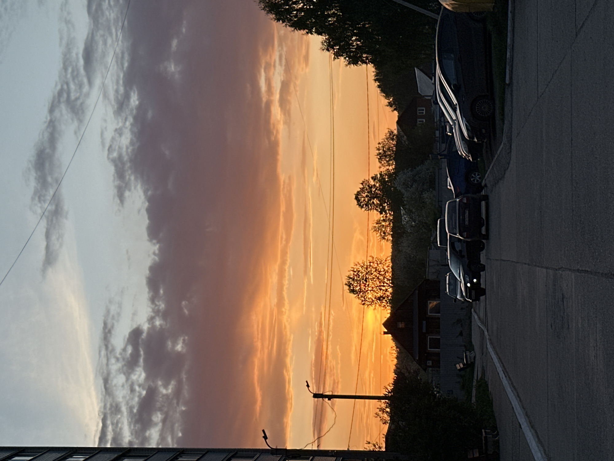 The image captures a sunset view from a street in Chaikovsky, Russia. The sky is filled with orange and pink clouds. Power lines stretch across the sky. On the right side of the image, there is a row of parked cars along the edge of a street. The street is asphalt and appears to be in a residential area. Houses are visible behind the cars and trees. Some trees are silhouetted against the sunset.