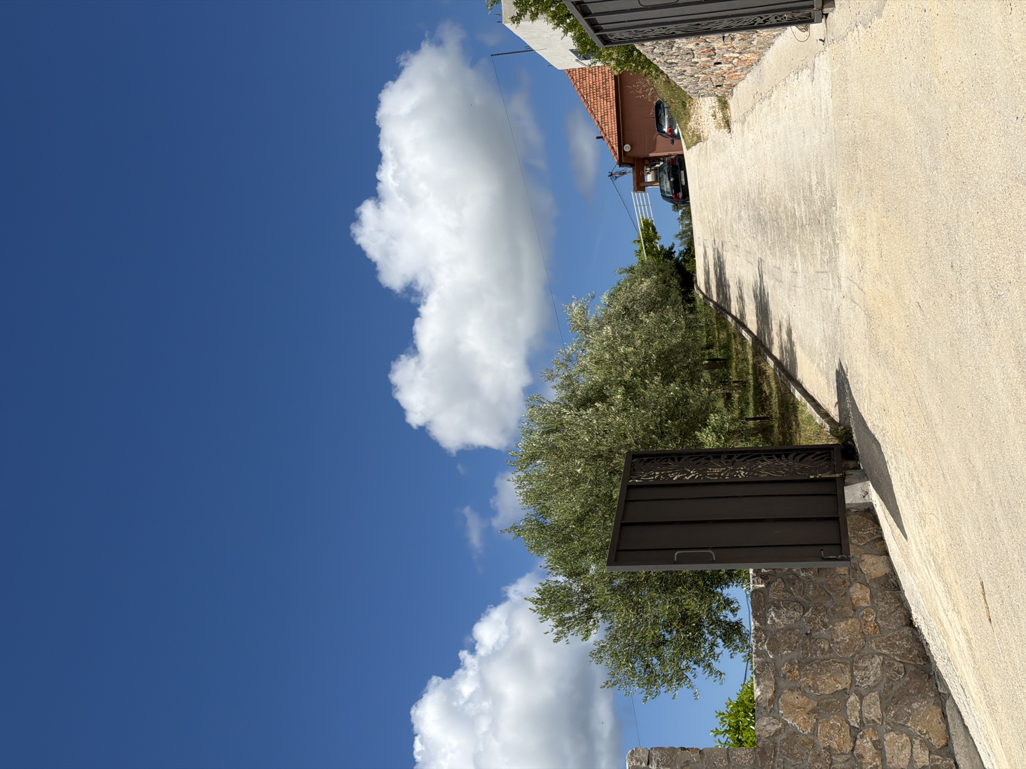 A sunlit outdoor scene unfolds, capturing a paved pathway bordered by lush greenery and stone walls in Bratešići, Montenegro. A vast, clear blue sky dominates the upper portion of the frame, dotted with large, fluffy white cumulus clouds.
The pathway, light-colored and likely concrete or gravel, extends diagonally from the foreground into the distance. On its left, a rustic stone wall rises, partially obscuring the lower sections of abundant green trees, possibly olive groves, that line the property. A dark metal gate, featuring decorative scrollwork on its upper panel, is positioned in the lower left, adjacent to the stone wall, suggesting an entrance. Long, distinct shadows of the trees are cast across the sun-drenched pathway, indicating a midday to early afternoon time.
In the background on the right, the warm terracotta-tiled roof of a building is visible, hinting at local architectural style. A dark-colored car is parked near this structure. Thin wires can be seen stretched across the bright sky near the building. The scene is devoid of people or explicit activity, presenting a tranquil and well-lit landscape under a brilliant blue sky, characteristic of a sunny day in the region. There is no visible text.