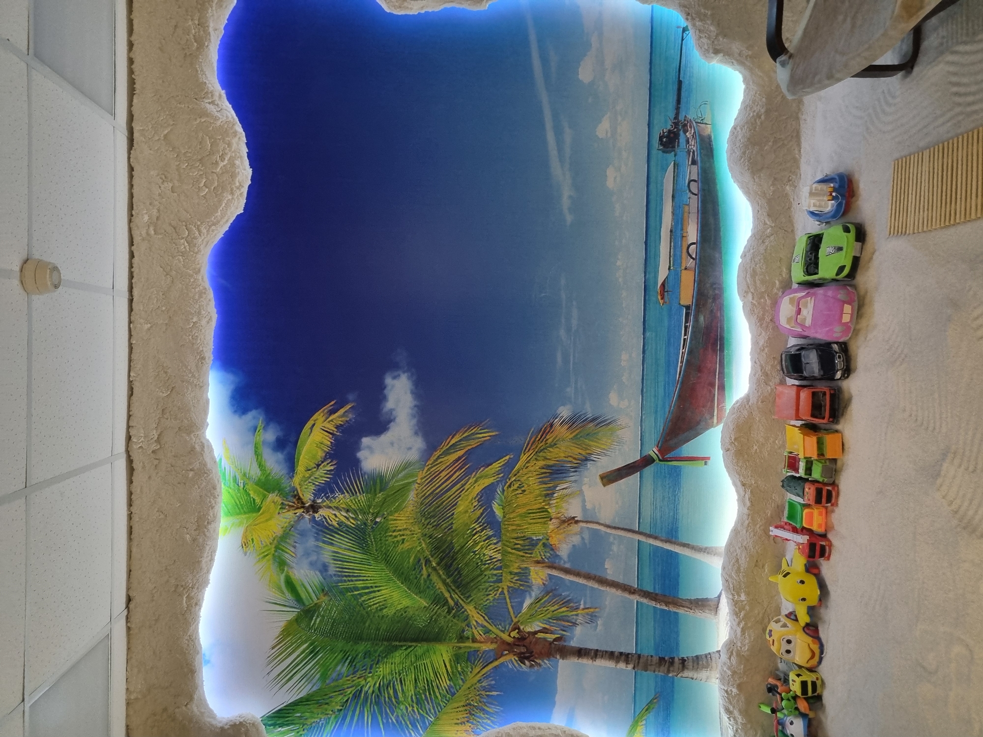 The image shows a ceiling with a beach-themed mural, possibly in a building in Chaikovsky, Russia. The mural includes a blue sky, clouds, palm trees, and a boat. The ceiling is surrounded by textured, white material. On the right side of the image are toy cars lined up on a surface. There is also a partially visible chair in the upper right corner and a rectangular wood feature. The rest of the ceiling appears to be standard white ceiling tiles.