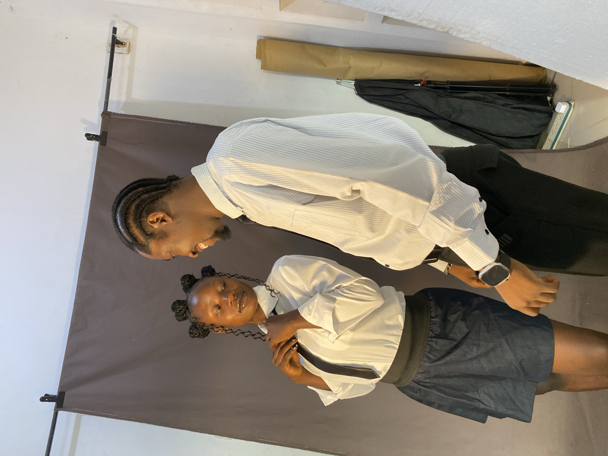 An indoor portrait photograph features two young Black individuals posing against a dark grey fabric backdrop. The setting appears to be a makeshift studio or a room adapted for photography, with a light-colored wall visible above the backdrop. In the background, rolled brown paper, a folded black umbrella or tripod, and a power strip are visible, along with textured white ceiling material.
The person on the left is taller, wearing a light-colored, vertically striped long-sleeved shirt and black trousers. Their hair is styled in neat braids. They are looking down and smiling towards the other person, their expression conveying warmth. A smartwatch is visible on their left wrist.
The person on the right is shorter, wearing a plain white long-sleeved shirt, a dark tie or ribbon, and dark blue shorts. Their braided hair is styled into two buns on the sides of their head. They are holding their tie with both hands and gazing slightly upwards with a subtle, serene expression. Their fingernails are painted dark.
Both individuals seem to be engaged in a casual photoshoot, with a friendly interaction evident between them. The overall mood is relaxed and pleasant. The image provides no specific time of day or weather indicators beyond being an indoor scene. This media was captured in Bukuru, Nigeria. No visible text is present in the image.