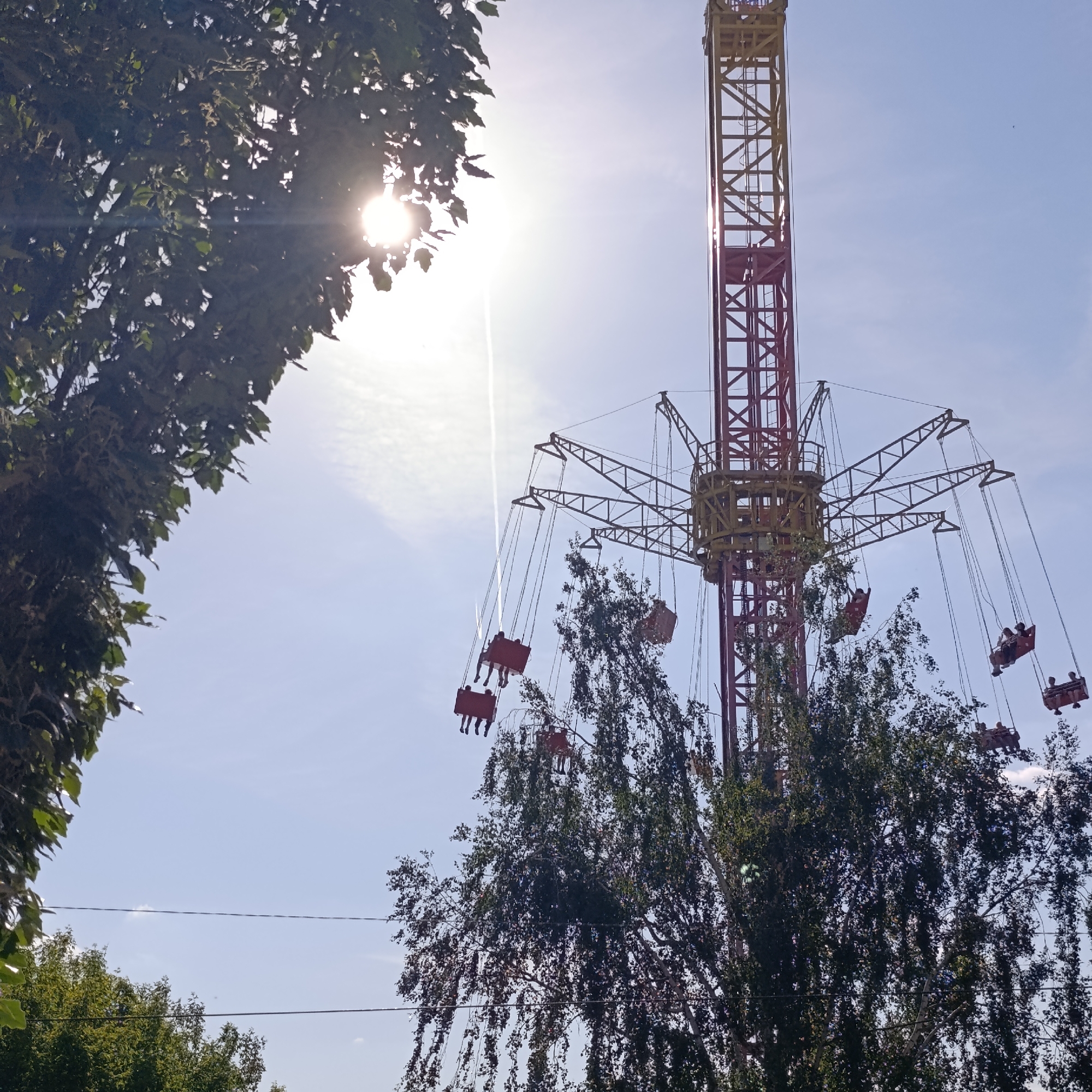 The image depicts a chair swing ride at an amusement park in Izhevsk, Russia. The ride features a tall, red and yellow tower with swing seats suspended from the top. The ride appears to be in operation, with several seats occupied by people. The background consists of a clear, bright sky, partially obscured by trees in the foreground. The sun is visible through the trees.