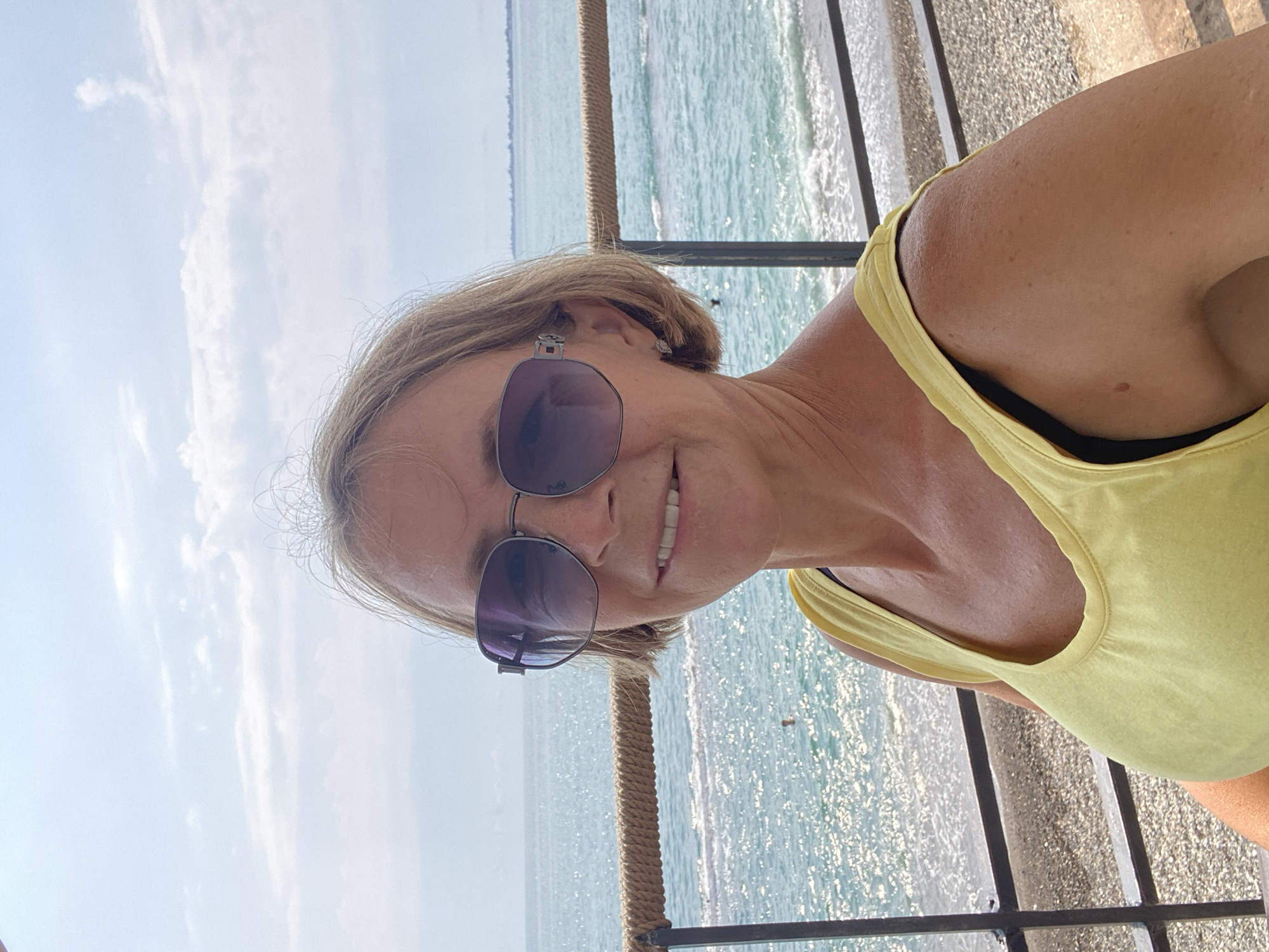 A woman with blonde hair and sunglasses is taking a selfie outdoors. She is wearing a yellow tank top. The background shows the sea with waves, a beach, and a cloudy sky. The woman appears to be on a balcony or promenade overlooking the sea in Chabkwara, Abkhazia.