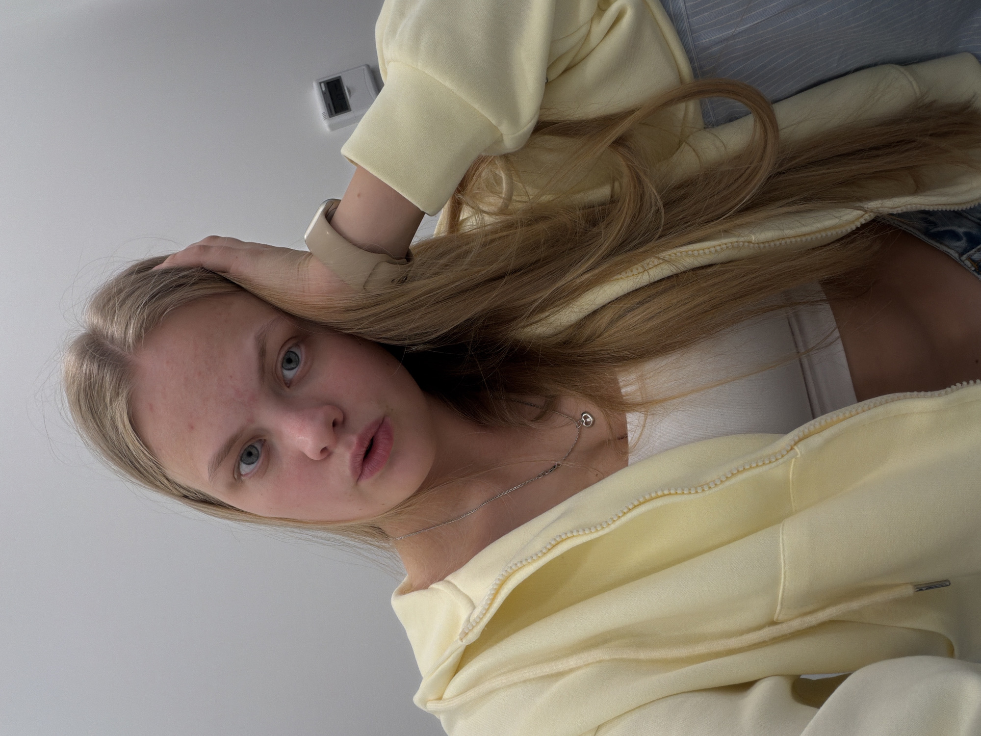 A young woman with fair skin and blue eyes looks directly at the camera, her left hand resting on her forehead. She has long, light brown hair that cascades over her left shoulder. She is wearing a white tank top and a pale yellow hooded sweatshirt. A thin silver necklace with a small pendant is visible on her neck. Her right wrist has a beige smartwatch. The background is a plain, light gray wall, with a small electronic device mounted on it in the upper left corner. The image appears to be taken indoors.
I cannot make any claims about the location.