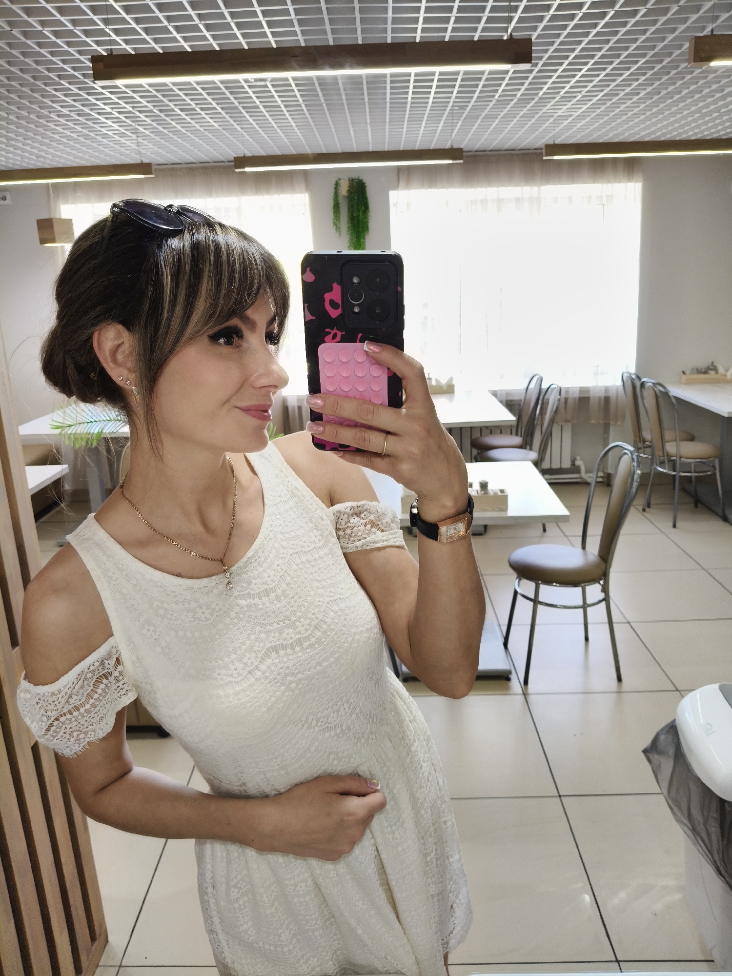A woman stands in what appears to be a cafeteria-style room, possibly in the city of Chaikovsky, Russia. She is holding up a phone, taking a selfie in a mirror. The woman is wearing a white lace dress with off-the-shoulder sleeves. She has brown hair with bangs and sunglasses on her head, and she is wearing jewelry, including a necklace and a watch. The background includes several tables and chairs, suggesting a dining area. The room has a drop ceiling with fluorescent lighting, and there's a window area covered with a sheer curtain. There also appears to be a plant in the background.