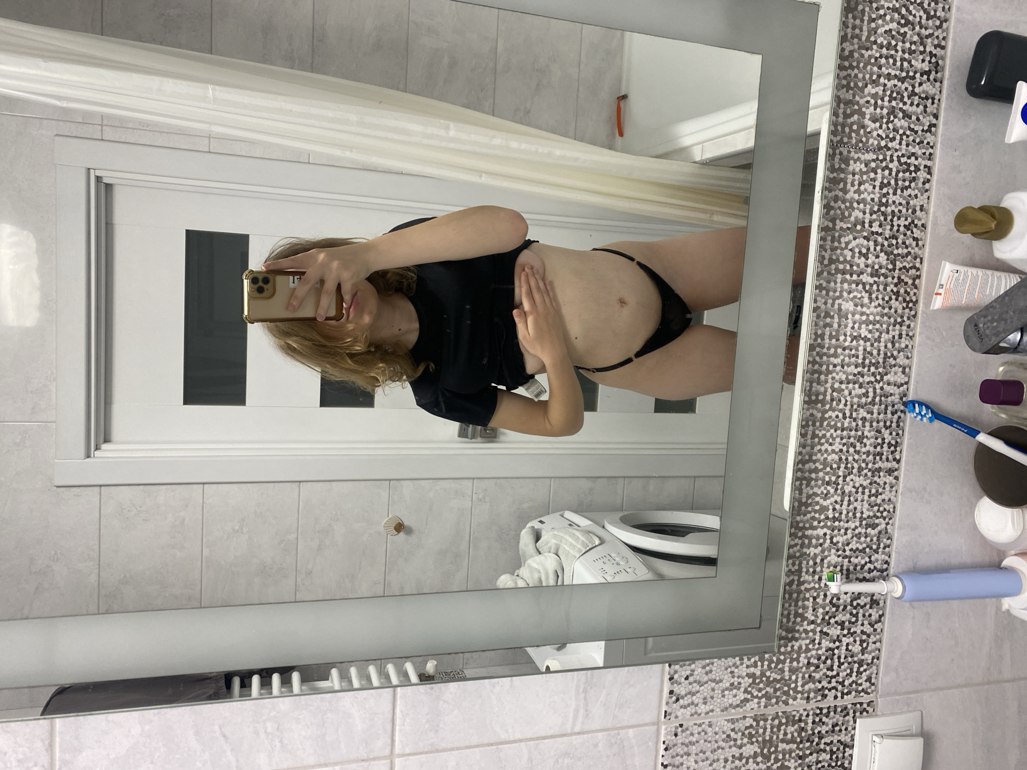 A mirror selfie captures a person in a modern bathroom, likely within a private residence in Ternopil, Ukraine. The subject, a individual with shoulder-length blonde hair, is seen from the chest down, holding a gold-cased smartphone that partially obscures their face. They wear a black short-sleeved top and black underwear, revealing their midriff with a navel piercing. Their left hand rests on their stomach, and a subtle smile is visible on their face.
The bathroom features light grey tiled walls, with a decorative mosaic-patterned tile strip running vertically beside the mirror. In the reflection behind the person, a white door, a shower curtain, a white toilet, and a washing machine with a visible control panel are discernable. An orange razor hangs on the tiled wall.
On the counter beside the mirror, various toiletries and personal care items are arranged: a dark bar of soap, a gold-capped bottle, tubes of cream (one possibly Nivea, another with "AVENTA" visible text), a faucet, a blue manual toothbrush, and a light blue electric toothbrush. The setting appears neat and well-maintained.